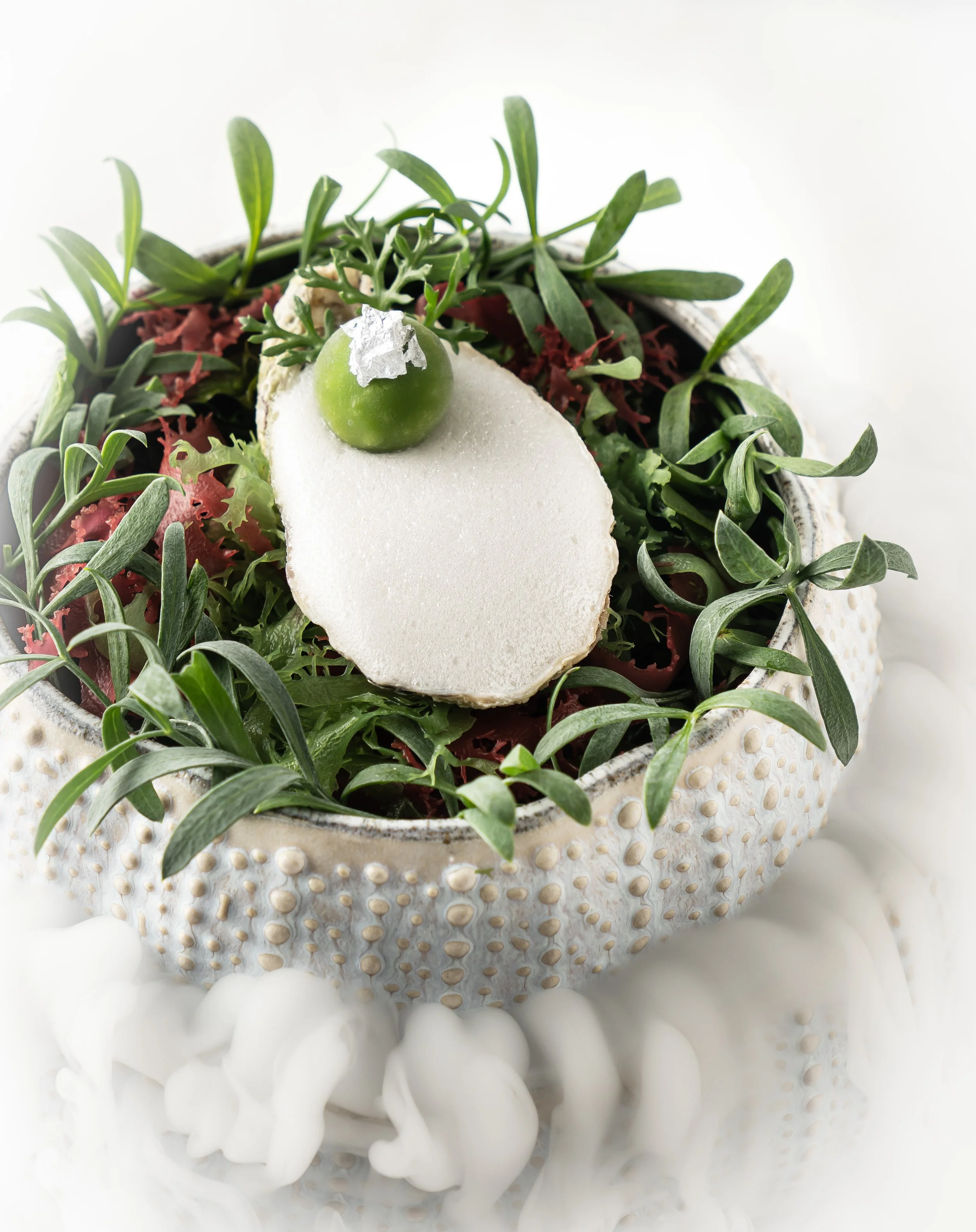 Decorative dish with green leaves, a white slice, and a small green sphere topped with silver garnish, surrounded by mist.