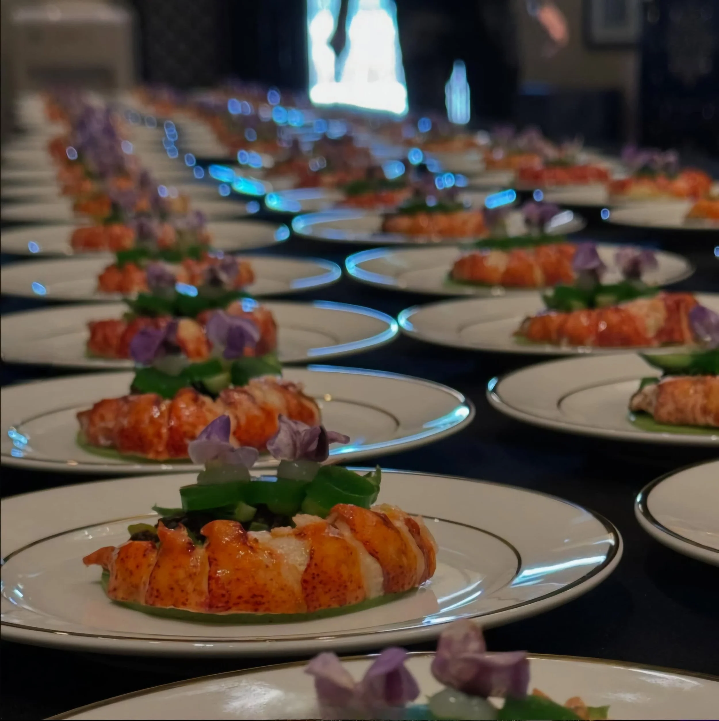 Rows of elegantly plated lobster dishes garnished with green vegetables and purple edible flowers.