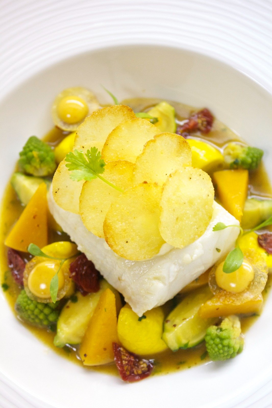 Gourmet fish dish with potato slices, vegetables, and garnish on a white plate.