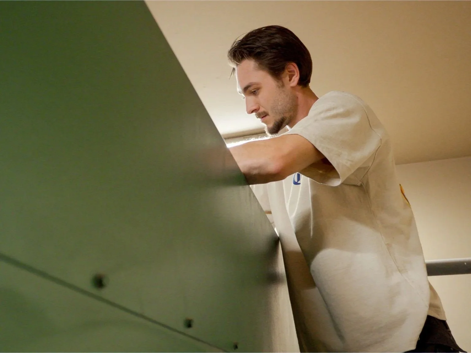 A young man with brown hair and a beard leaning over a green surface, focused and looking down at something, in a room with a beige ceiling.