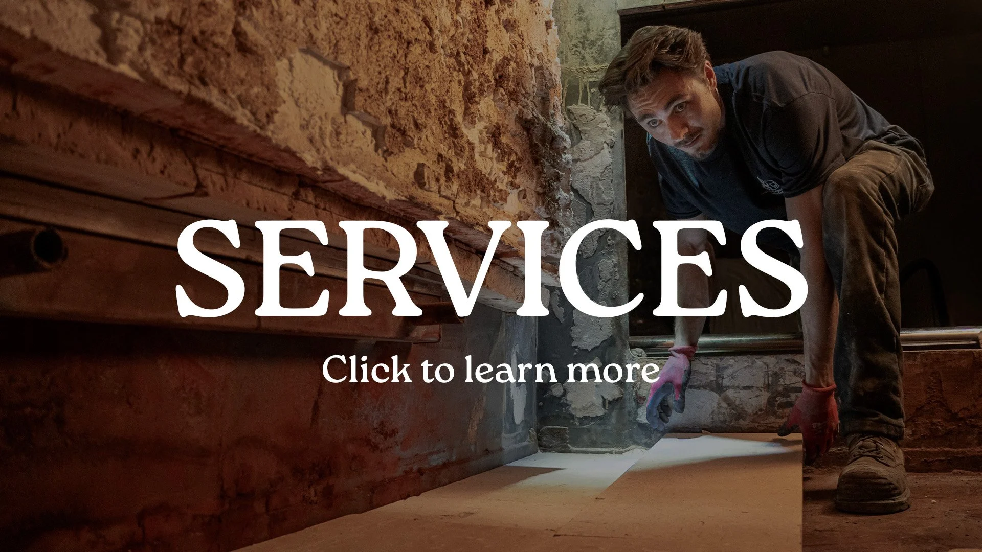 A man working on a construction site, crouched down near a brick wall, wearing a black shirt, brown pants, and gloves, with the word 'SERVICES' overlayed in large white letters and a call to action to click to learn more.