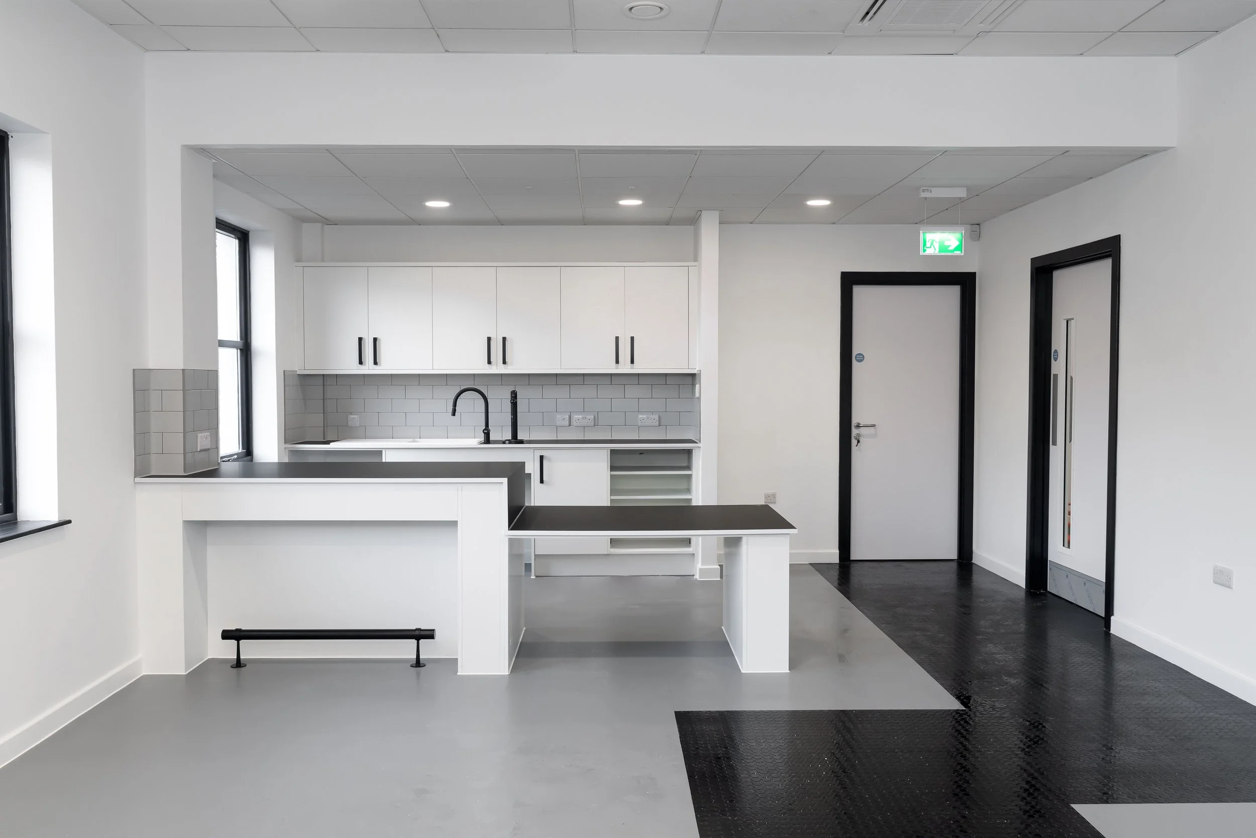 Modern, empty kitchenette with white cabinets, black handles, gray subway tile backsplash, black faucet, black countertop, gray flooring, black framed door, and an emergency exit sign, taken by an interior photography Edinburgh service.