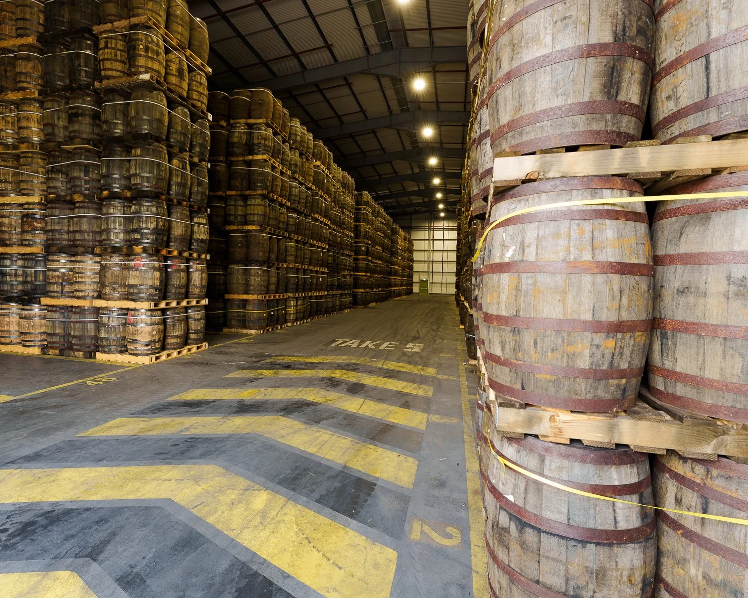 Warehouse with stacks of wooden barrels on pallets and a cleared aisle marked ‘TAKE 3,’ photographed by an industrial photography Glasgow service.