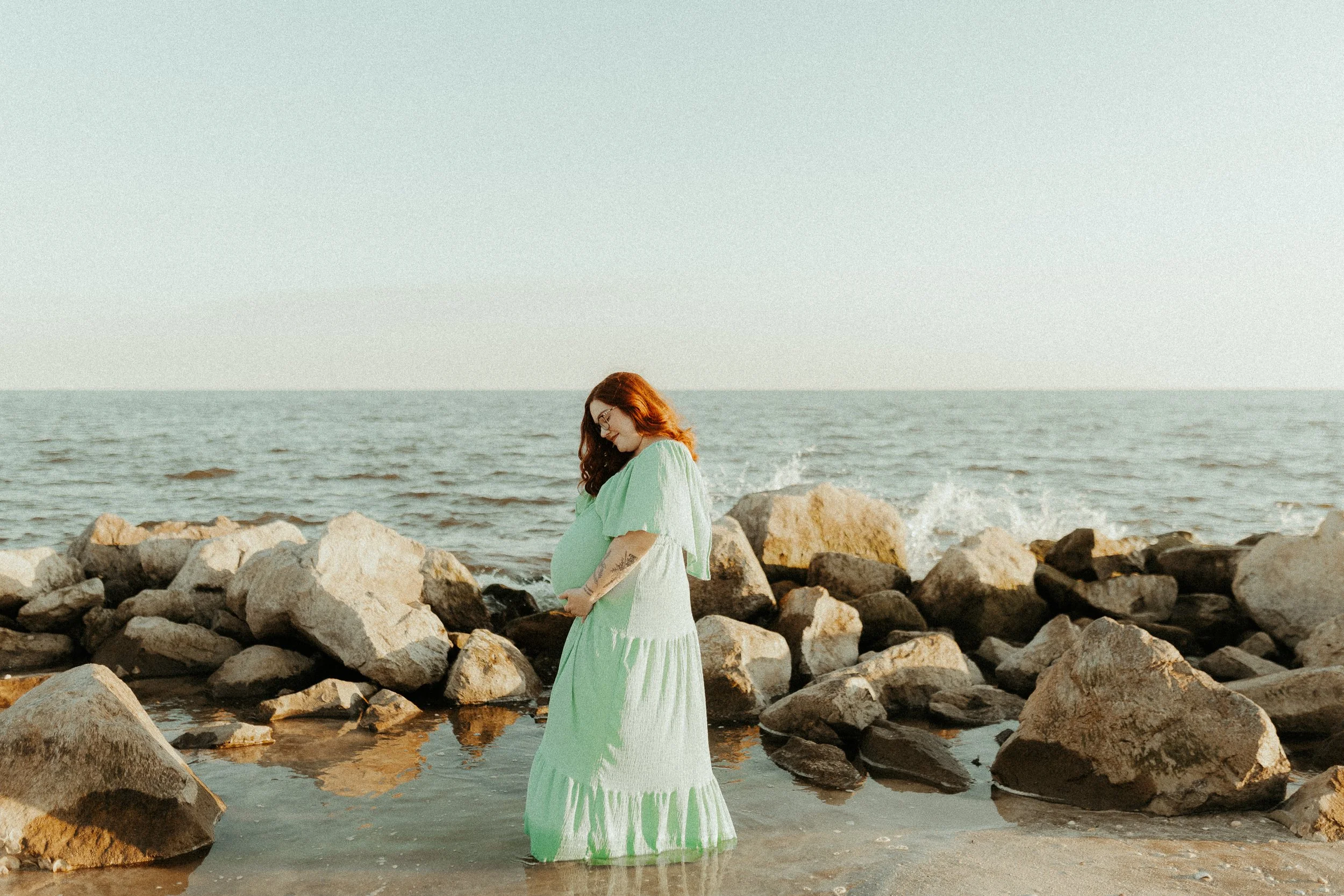 A pregnant woman in a light green dress standing on rocks by the ocean, looking down and cradling her belly.
