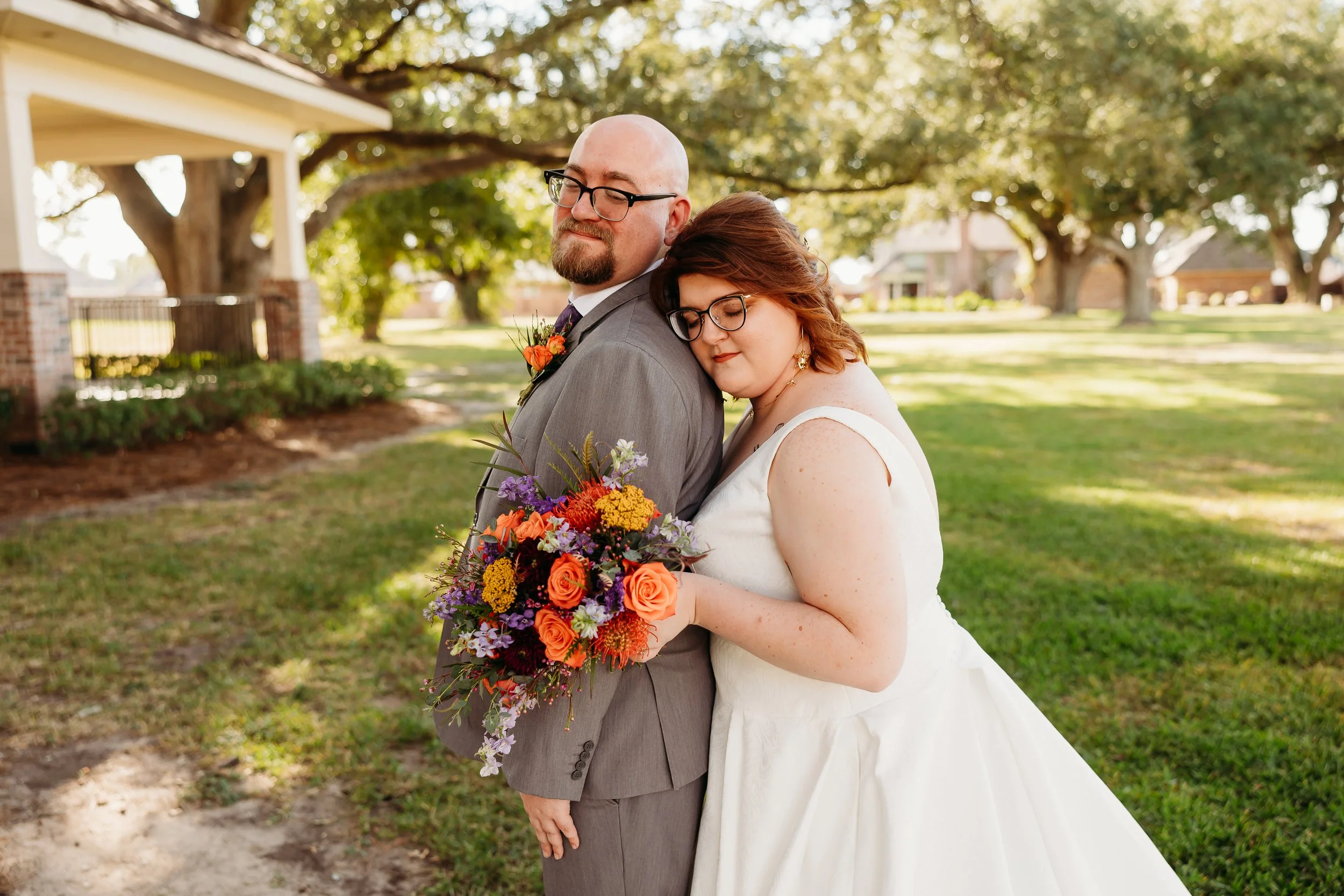 A bride and groom standing outdoors in a park with trees in the background, the bride resting her head on the groom's shoulder, holding a colorful bouquet of flowers, both with their eyes closed.