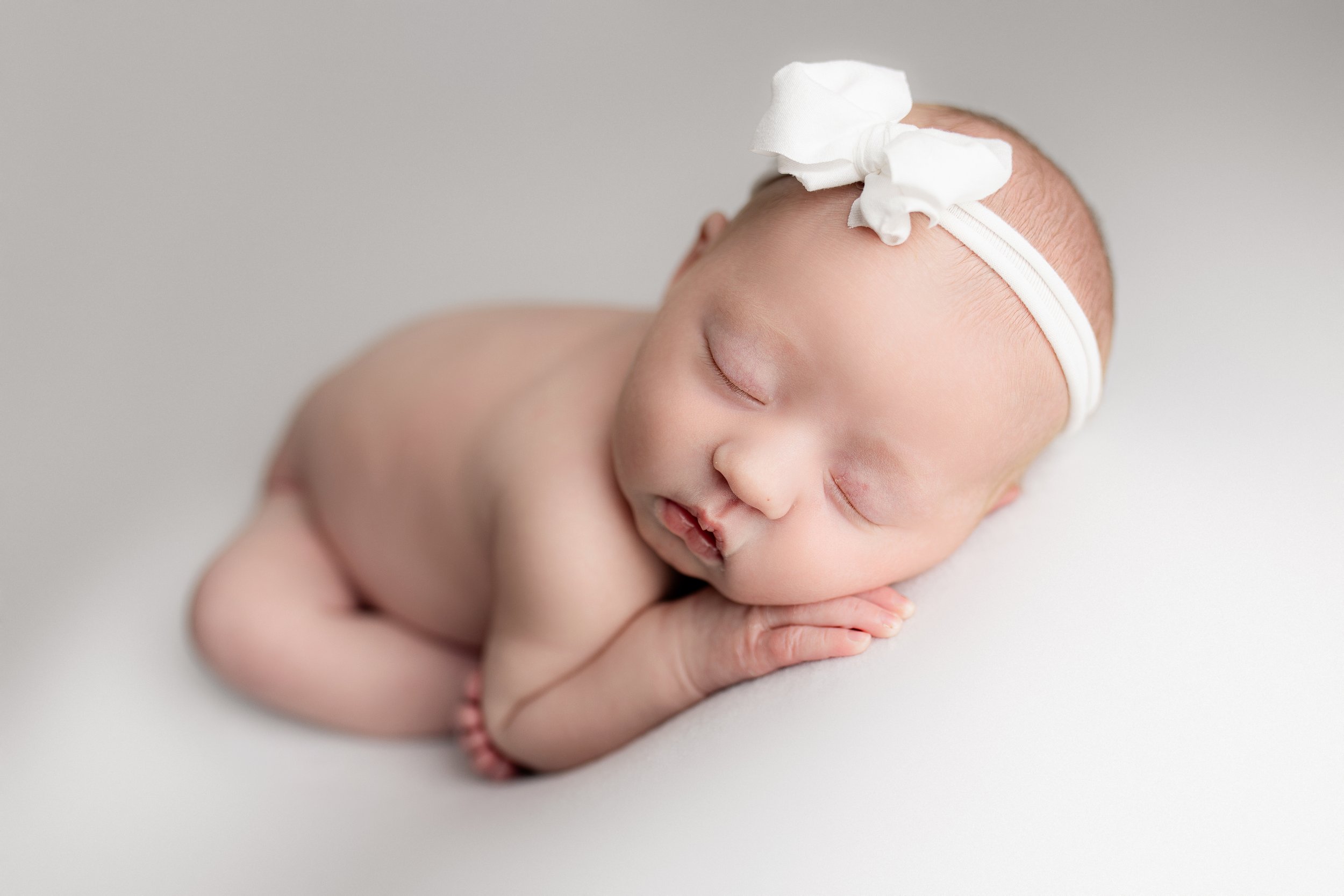 A sleeping newborn baby lying on a white surface, wearing a white headband with a bow.