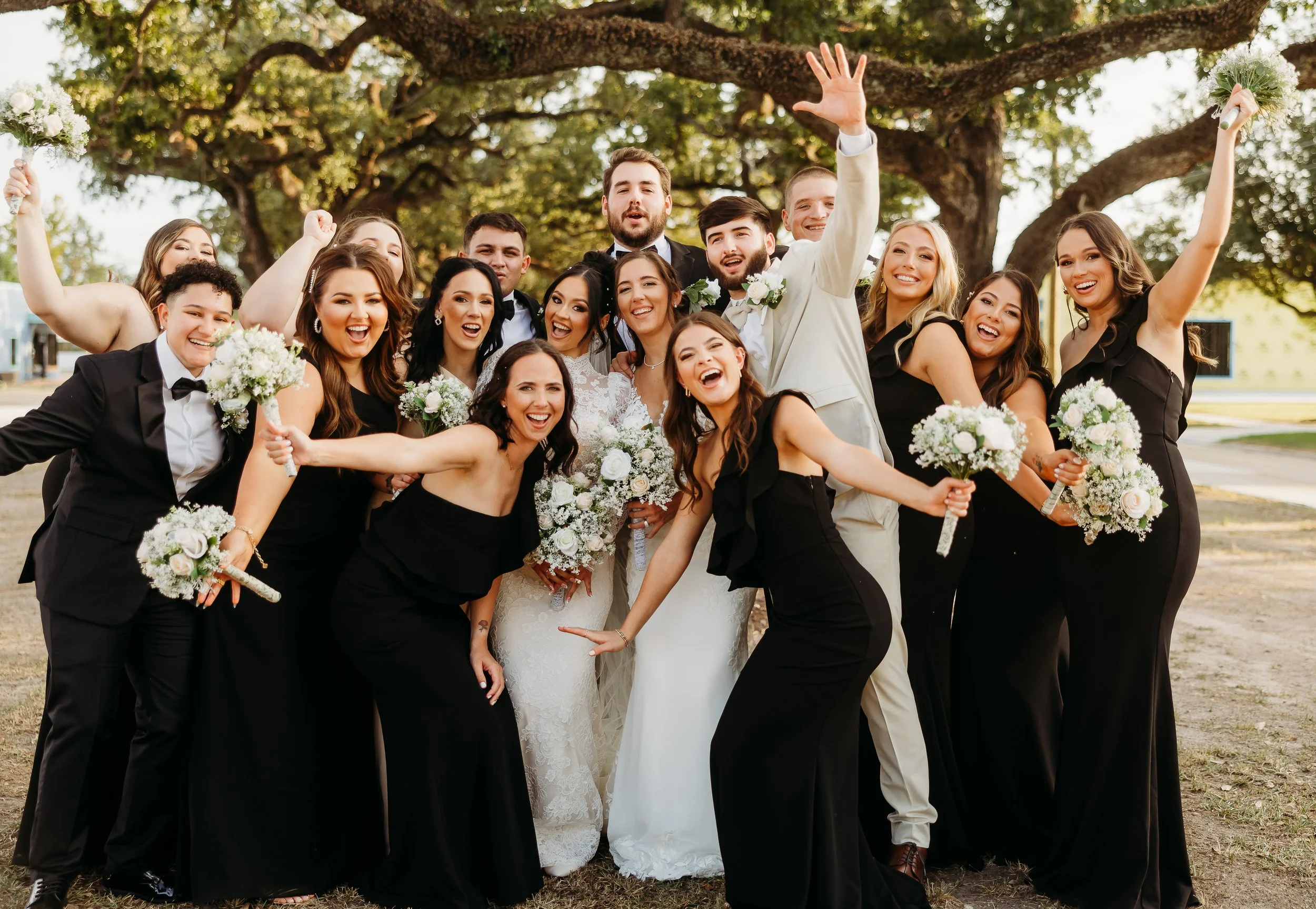 A group of wedding party members, including the bride, groom, bridesmaids, and groomsmen, posing outdoors under a large tree, celebrating with smiles and laughter, holding bouquets of white flowers.