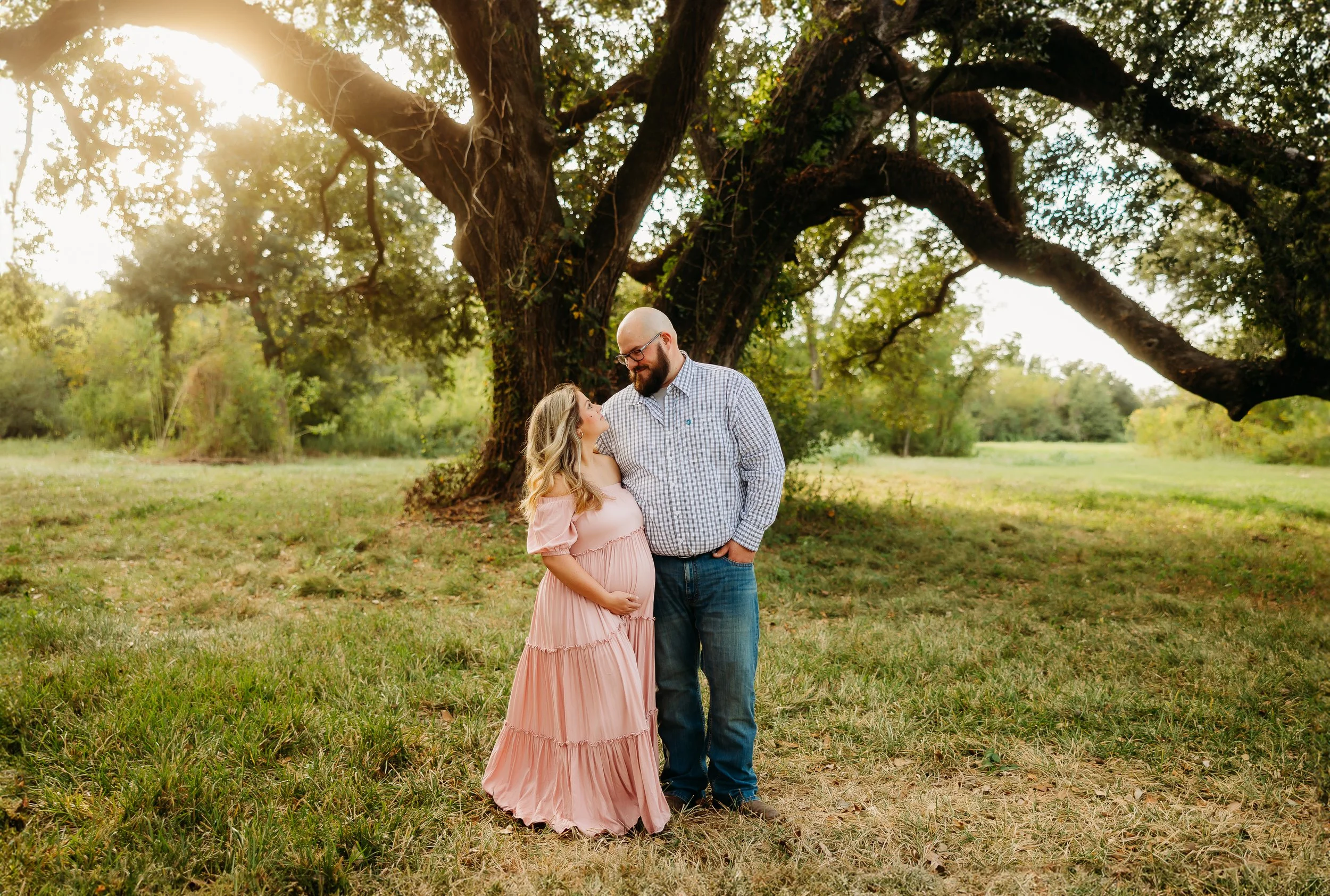 A pregnant woman and a man standing under a large tree, smiling and looking at each other, in a grassy field during sunset.