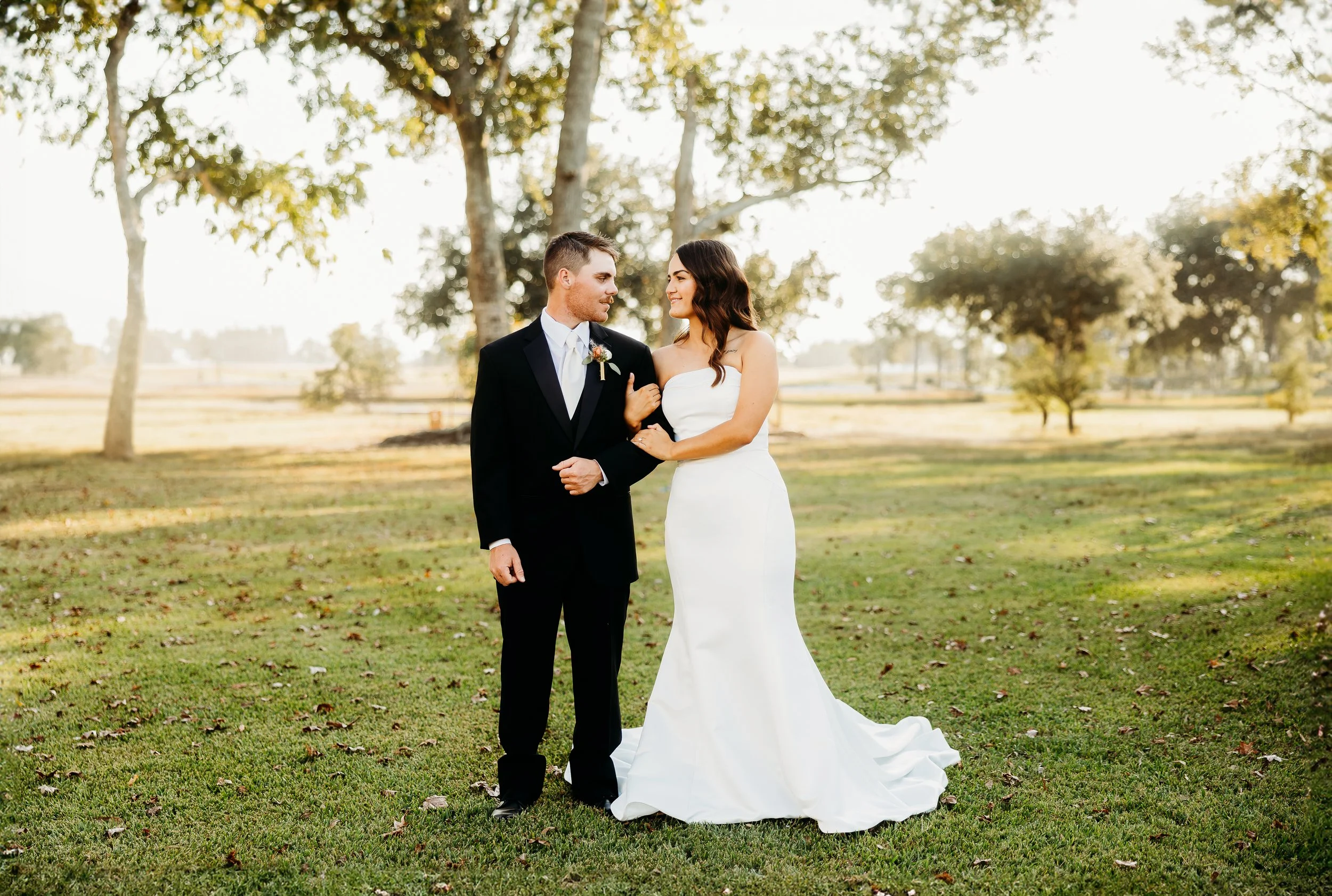 A newlywed couple standing on grass in a park with trees, smiling and looking at each other. The groom is wearing a black tuxedo with a white shirt and tie, and the bride is in a strapless white wedding dress. The scene is lit by natural sunlight.
