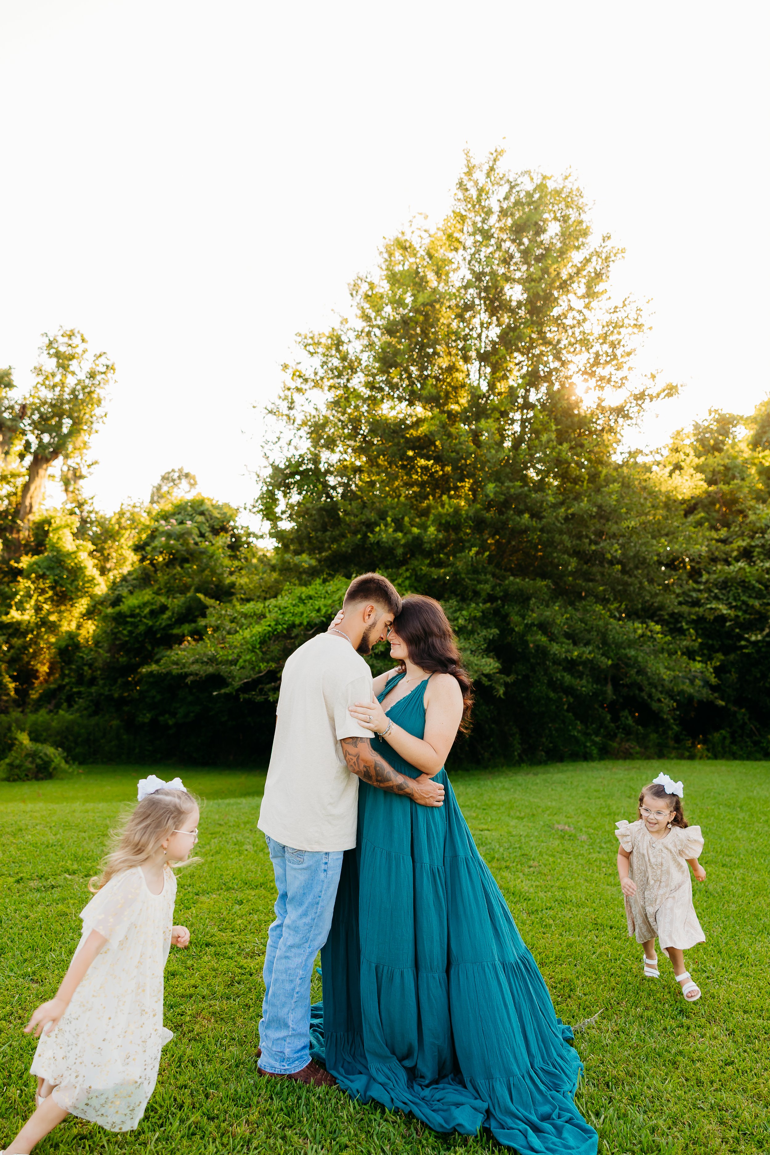 Four children, two boys and two girls, smiling and laughing outdoors during sunset, by a lake with trees in the background.