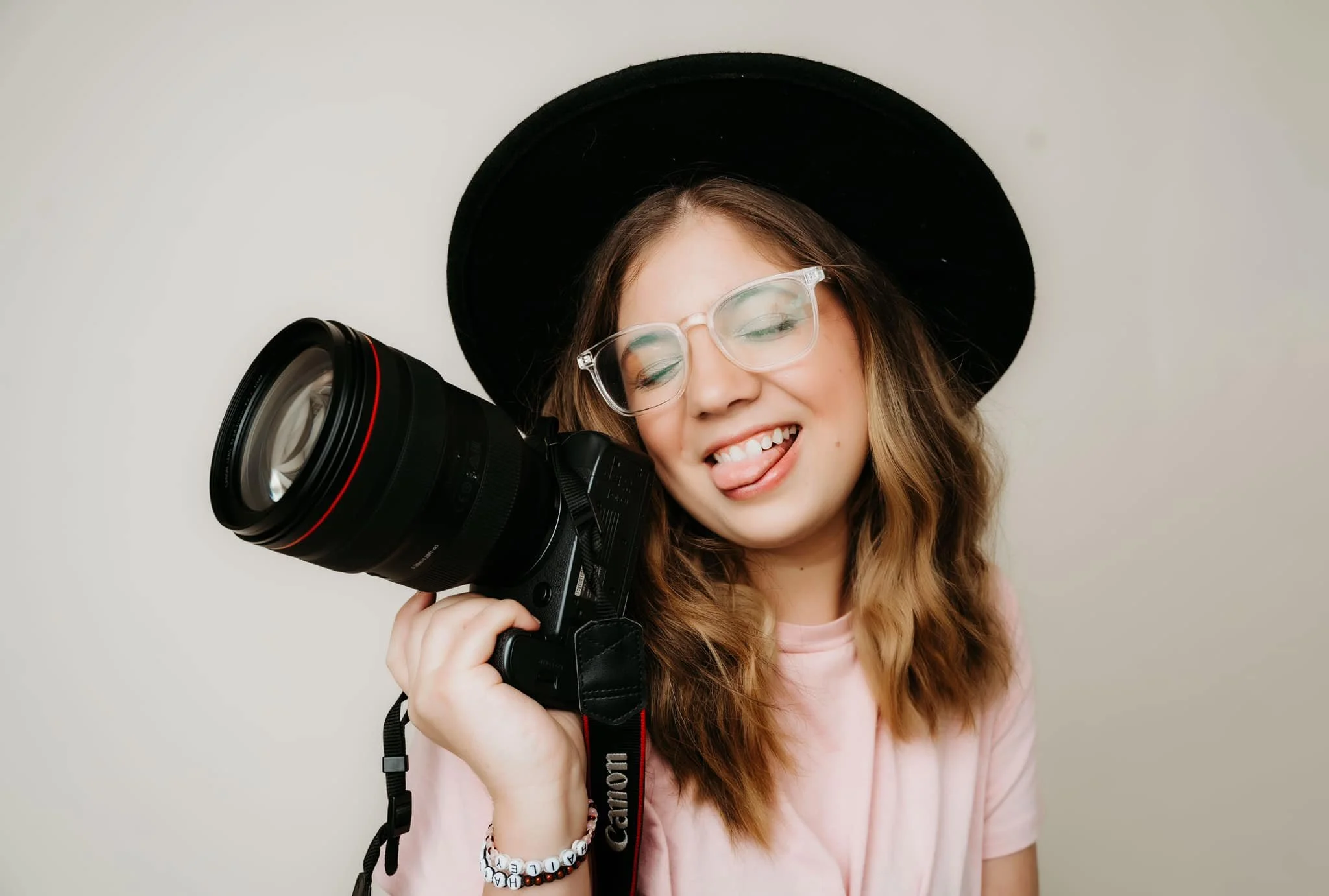 Young woman with glasses and a black hat making a playful face, holding a professional camera with a large lens against a plain background.