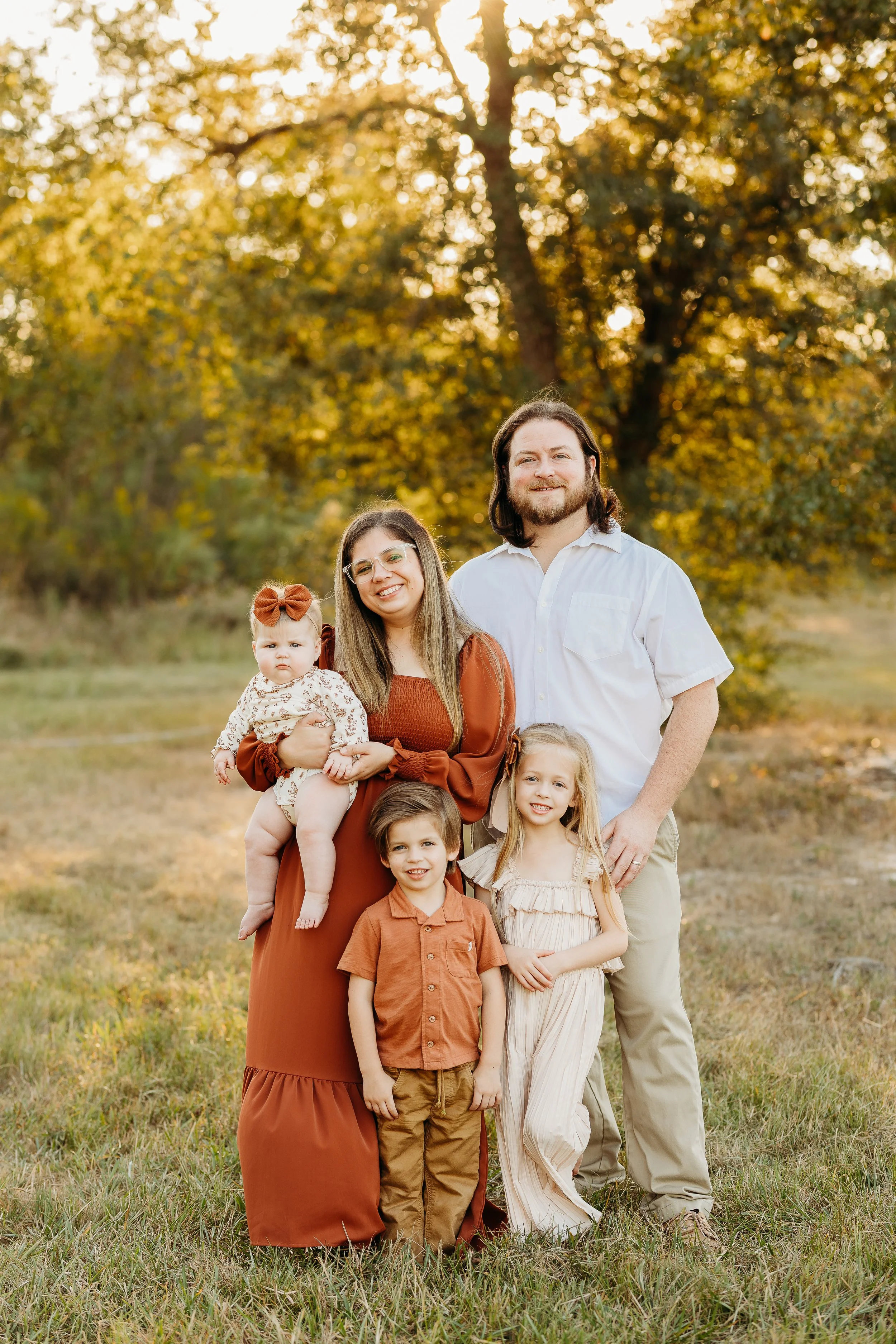 A family of five standing outdoors in a grassy area with trees in the background during fall, smiling at the camera.