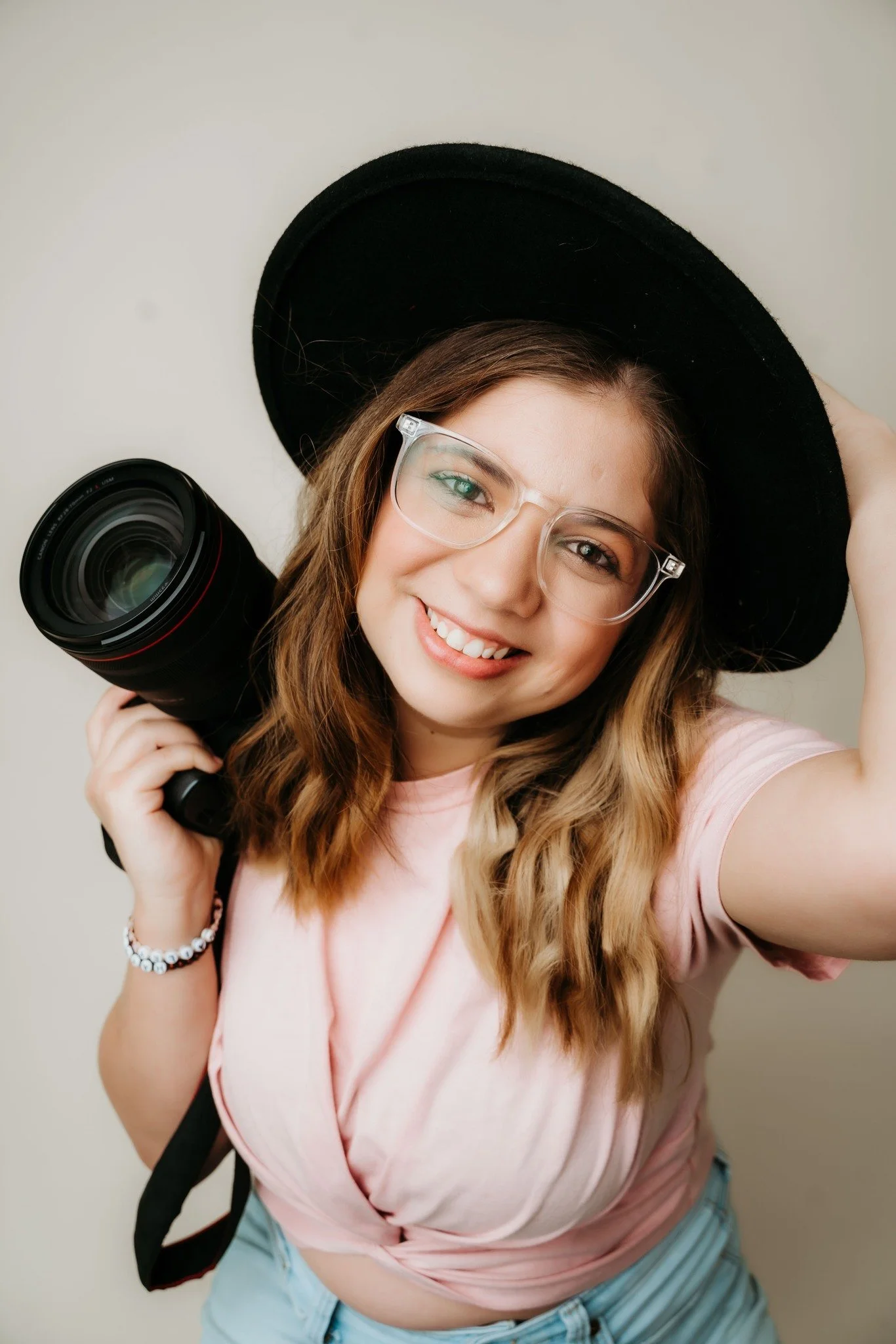 A young woman with wavy brown hair, wearing glasses, a black hat, a light pink top, and light blue jeans, smiling and holding a camera over her shoulder.