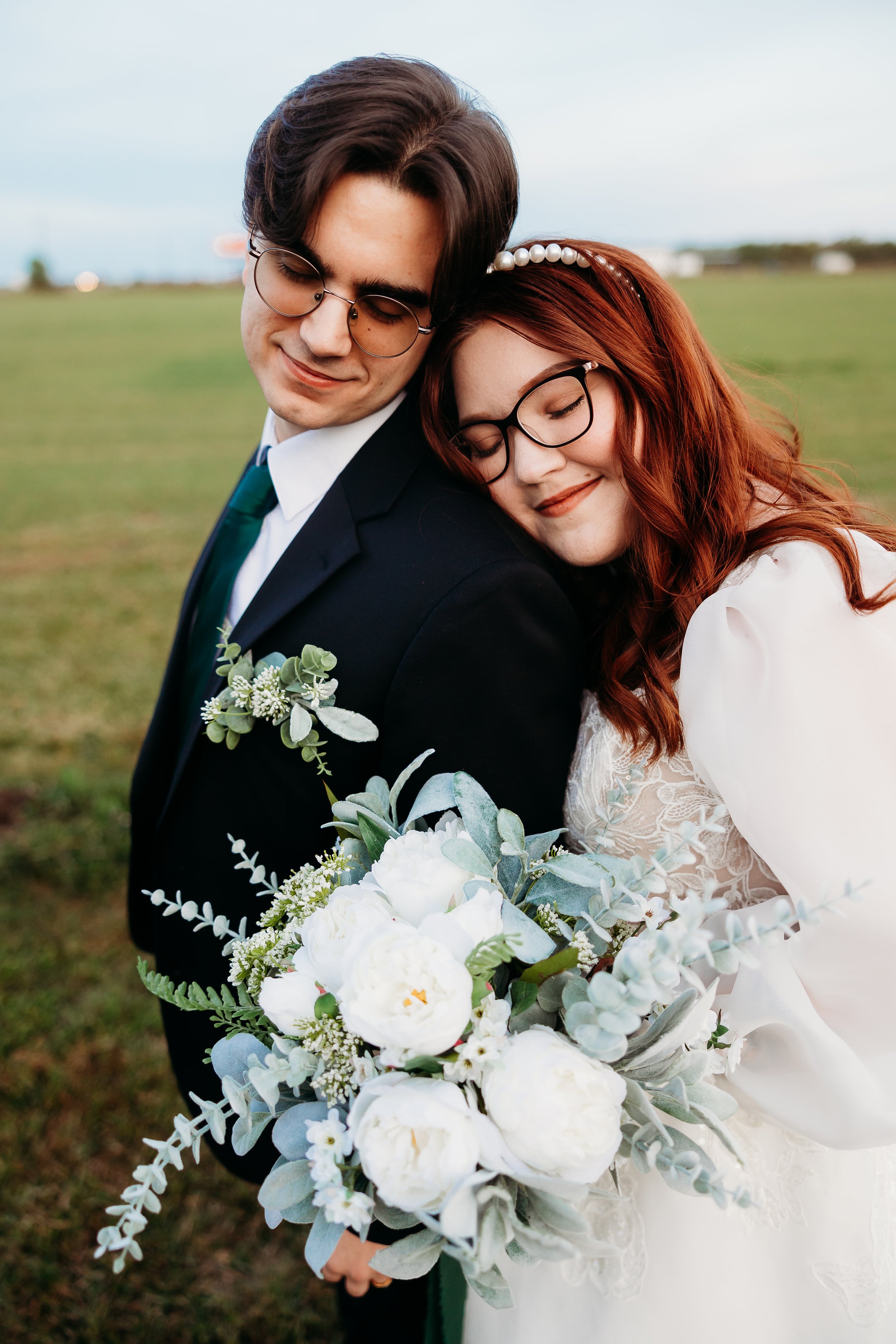 A newlywed couple embracing outdoors, with the bride holding a bouquet of white flowers and greenery, both with closed eyes and smiling softly.