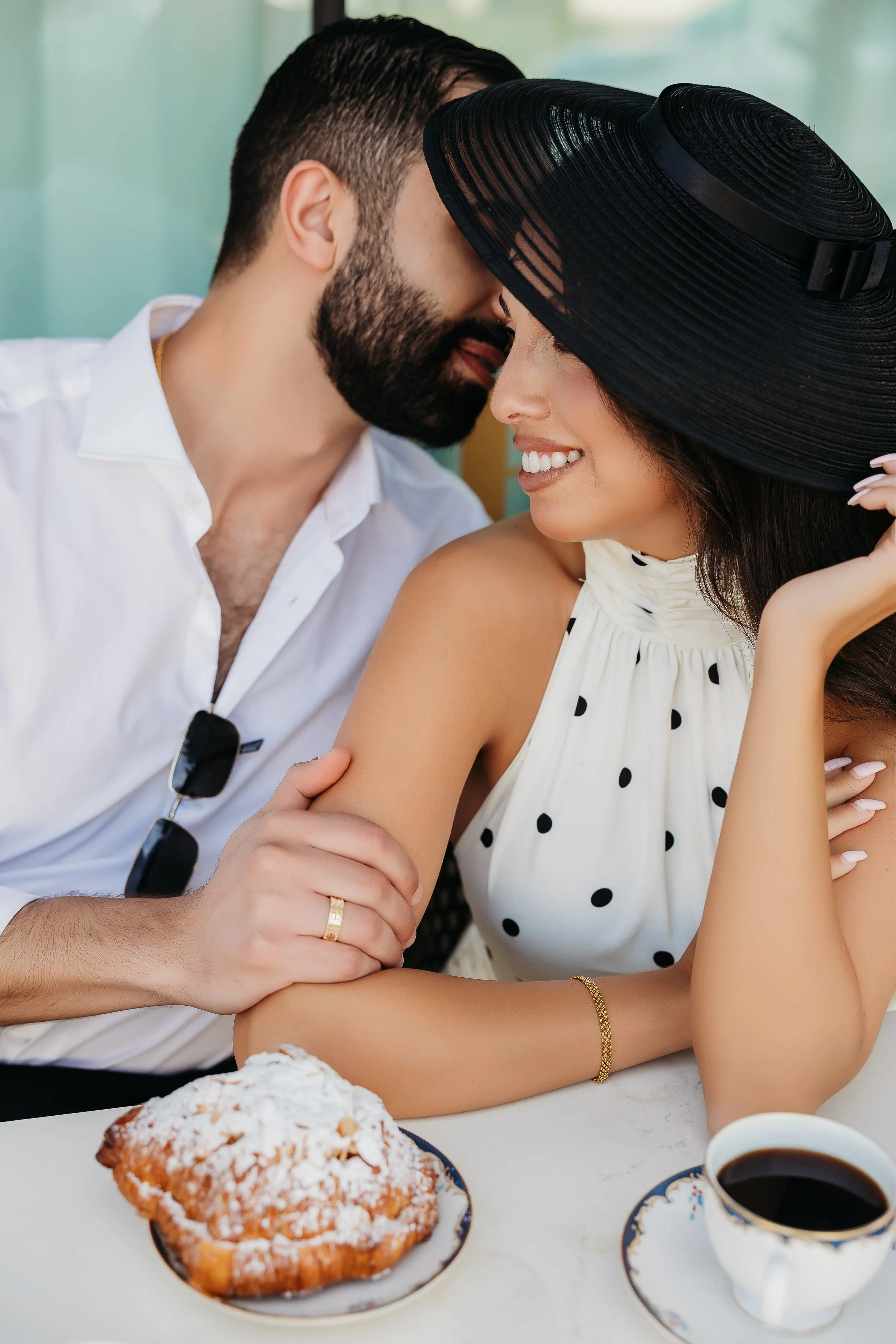 A couple sitting at a table, leaning close to each other, with the man whispering into the woman's ear. The woman is wearing a wide-brimmed black hat and a white dress with black polka dots, smiling happily. The table has a large cinnamon pastry, a c