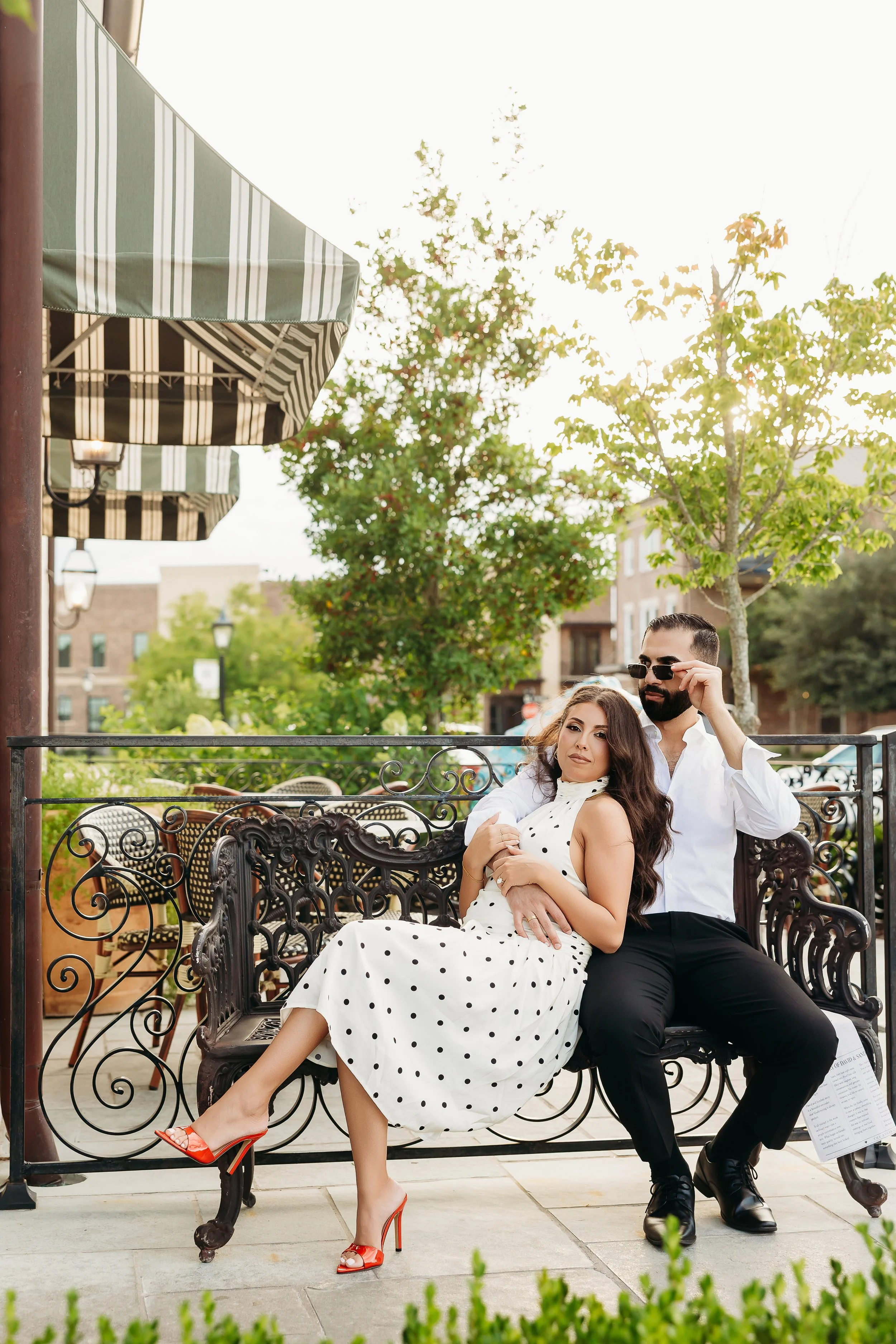 A woman sitting on a decorative black iron bench, wearing a white polka dot dress and high heels, with a man standing behind her, adjusting his sunglasses, in an outdoor cafe setting with trees and striped awnings.