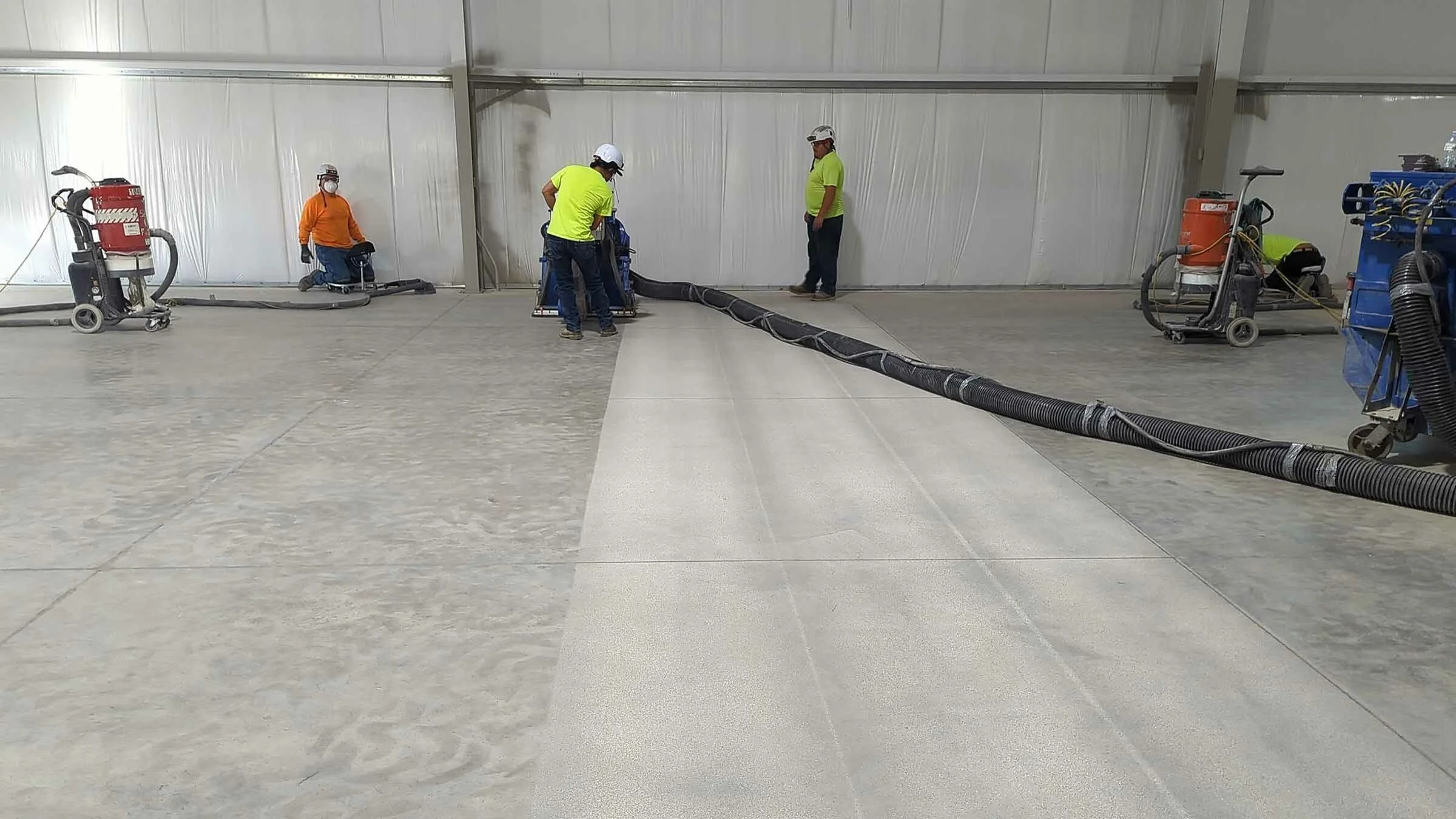 Workers installing flooring in a large industrial space, with some applying a floor coating and others working with large hoses and equipment.