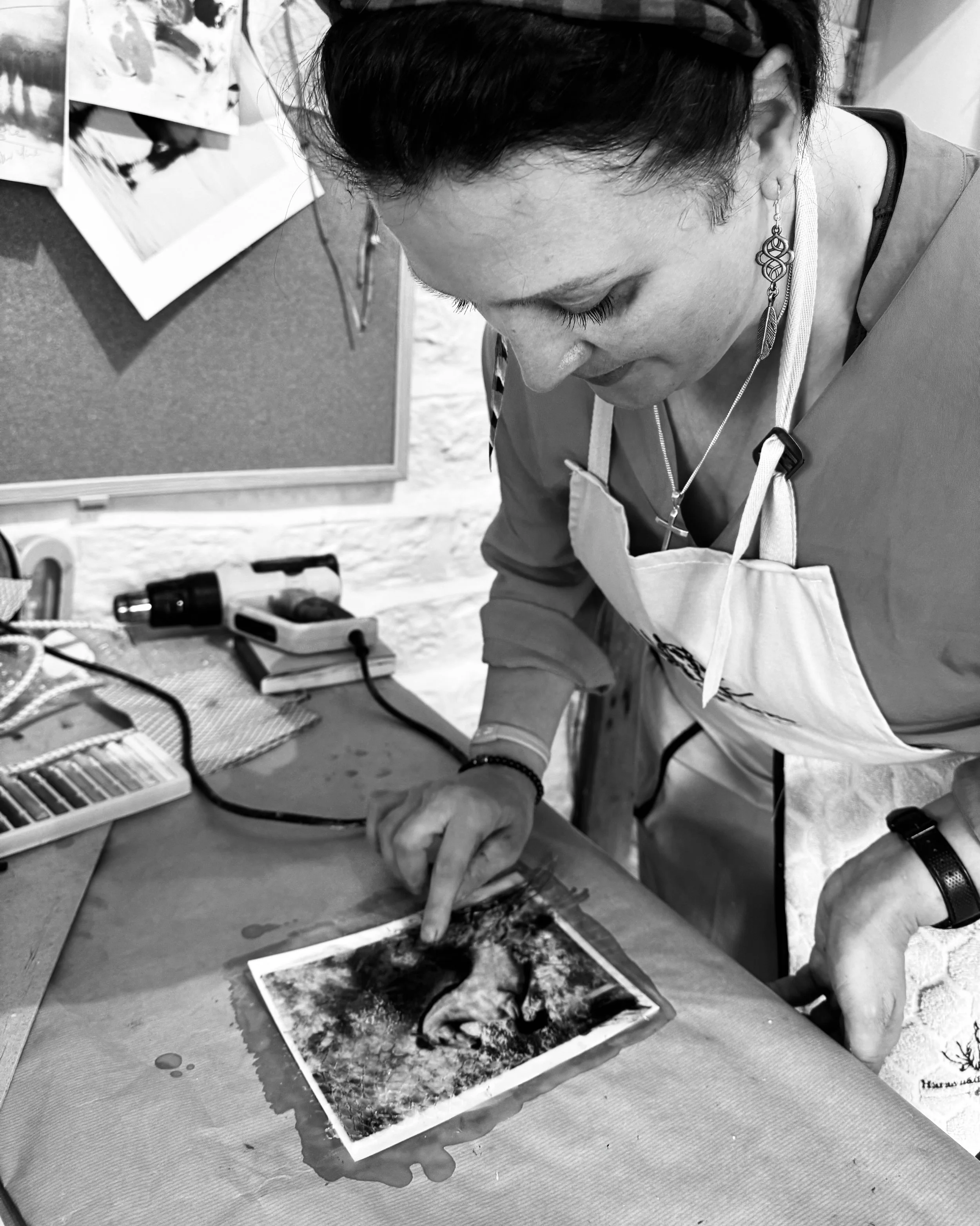 A woman with dark hair looking down as she points at a black and white photograph on a table, surrounded by art supplies and a corkboard with pinned photos in the background.