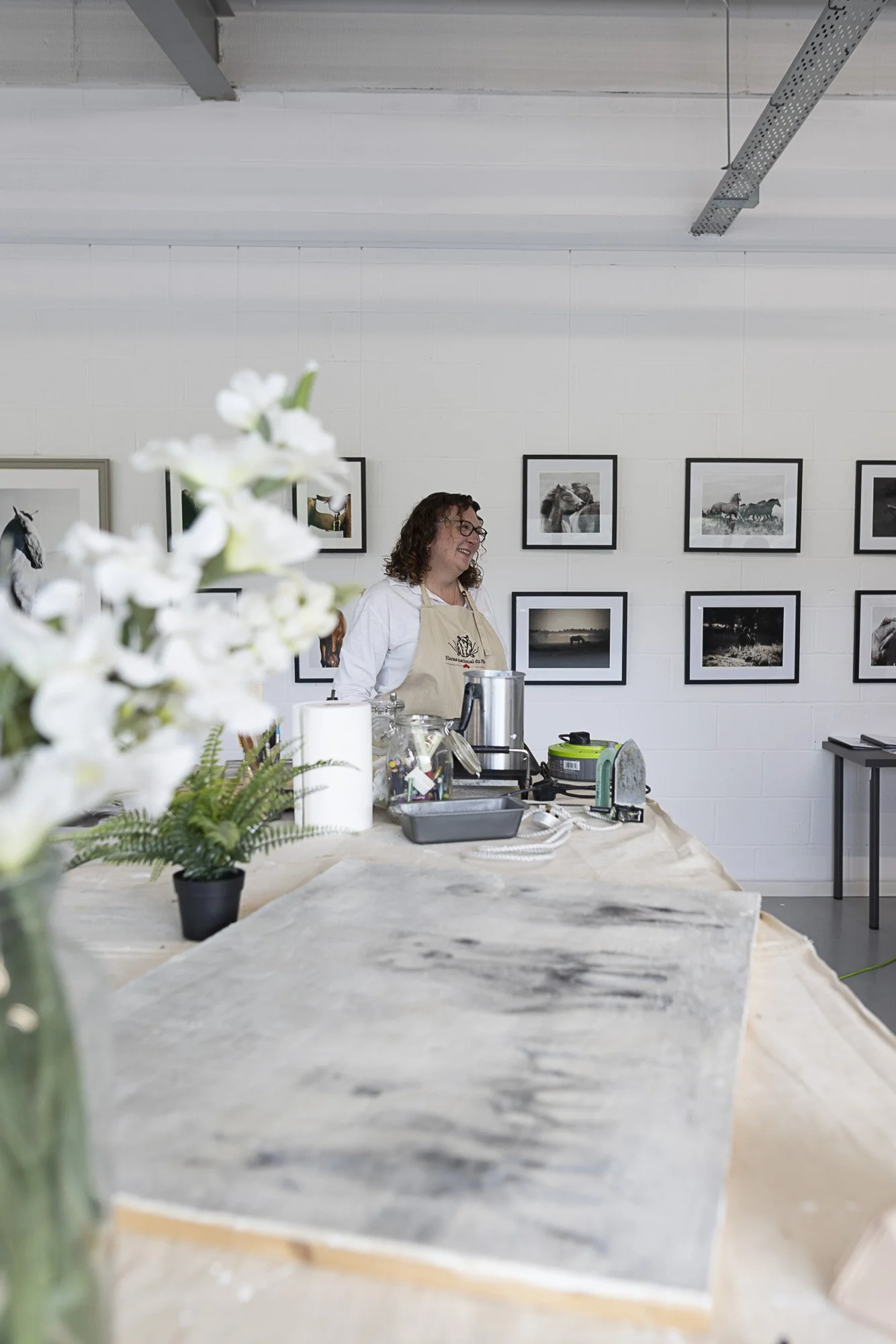 A woman with glasses and curly hair smiling, standing behind a table with art supplies in an art studio, with framed horse photographs on the wall in the background.