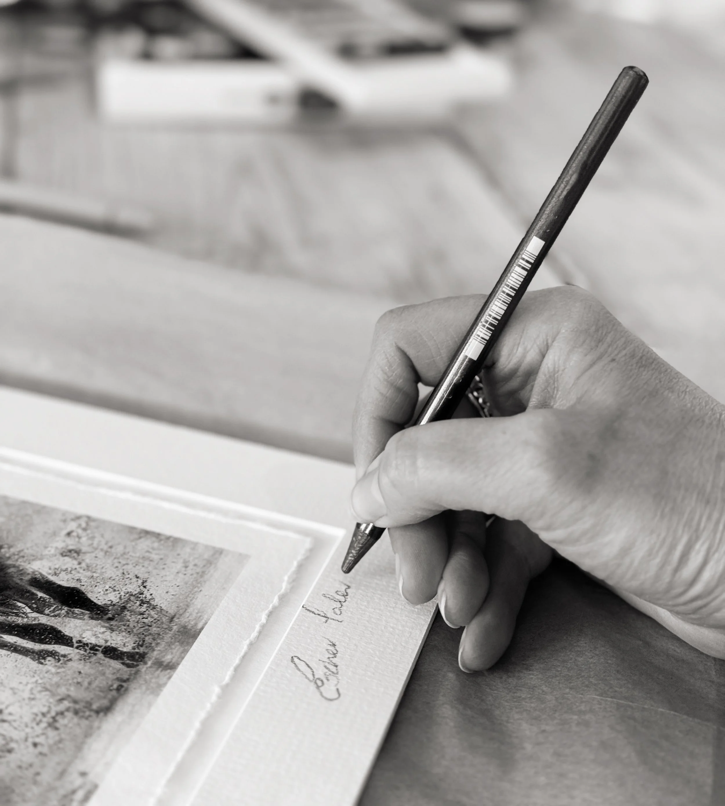 A hand writing 'Cherished Je' on a piece of paper with a pen, next to a framed photograph, in black and white.