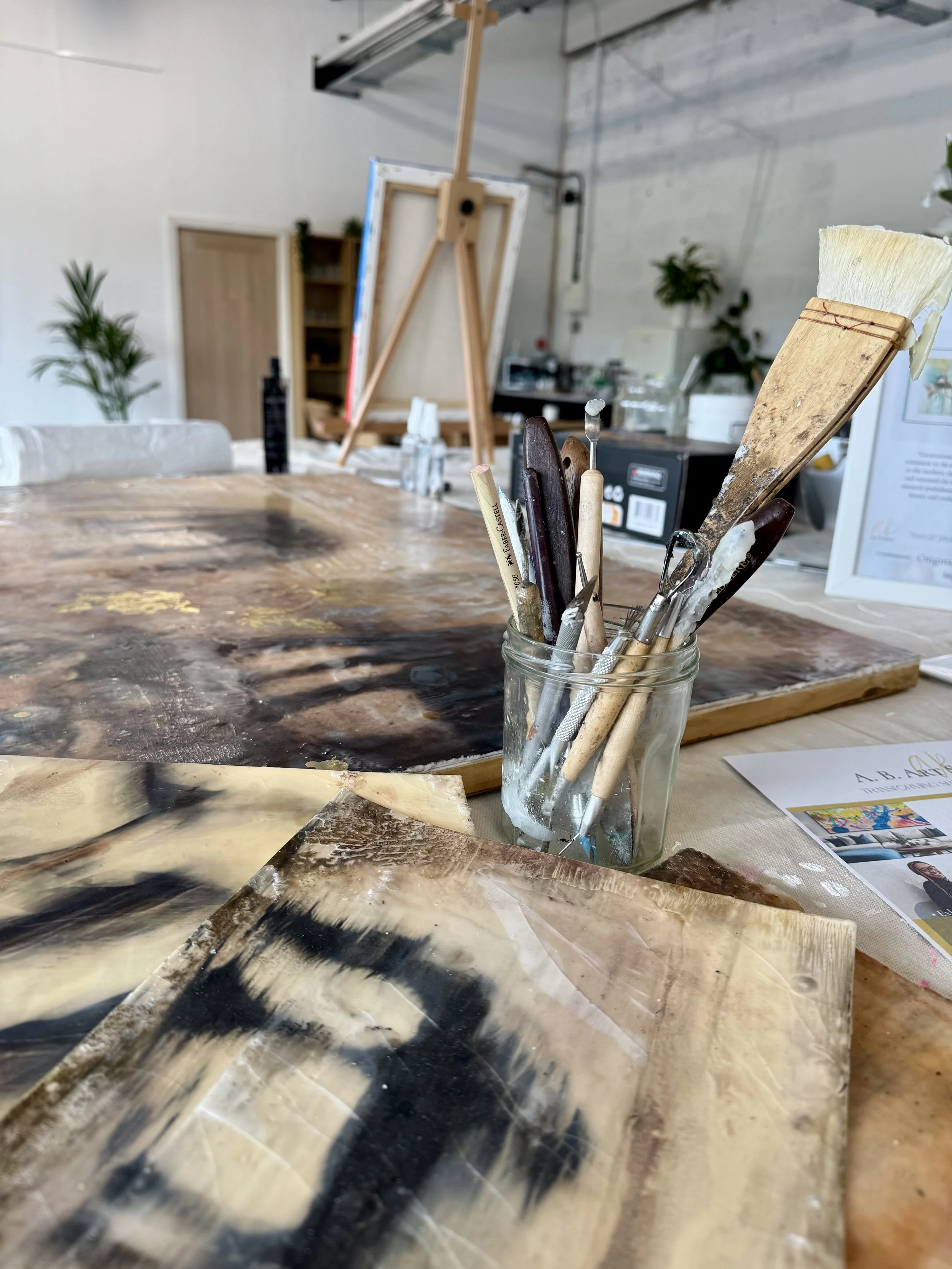 A workspace with paintbrushes in a glass jar, encaustic paintings, an easel, and various art supplies in an art studio.