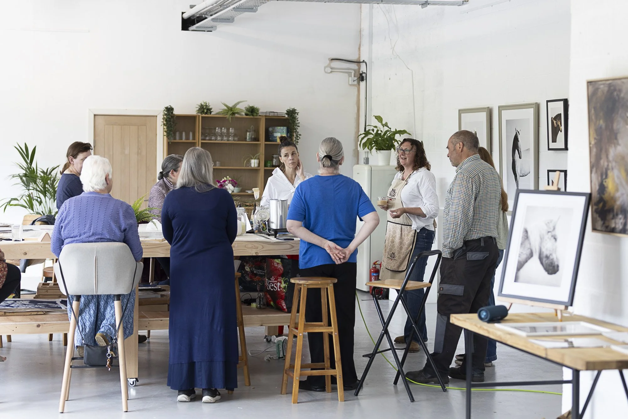 A group of people gathered in an art studio or gallery, listening to a woman with glasses who appears to be speaking or giving a presentation. The studio has framed animal artwork on the walls and tables with art supplies and photographs.