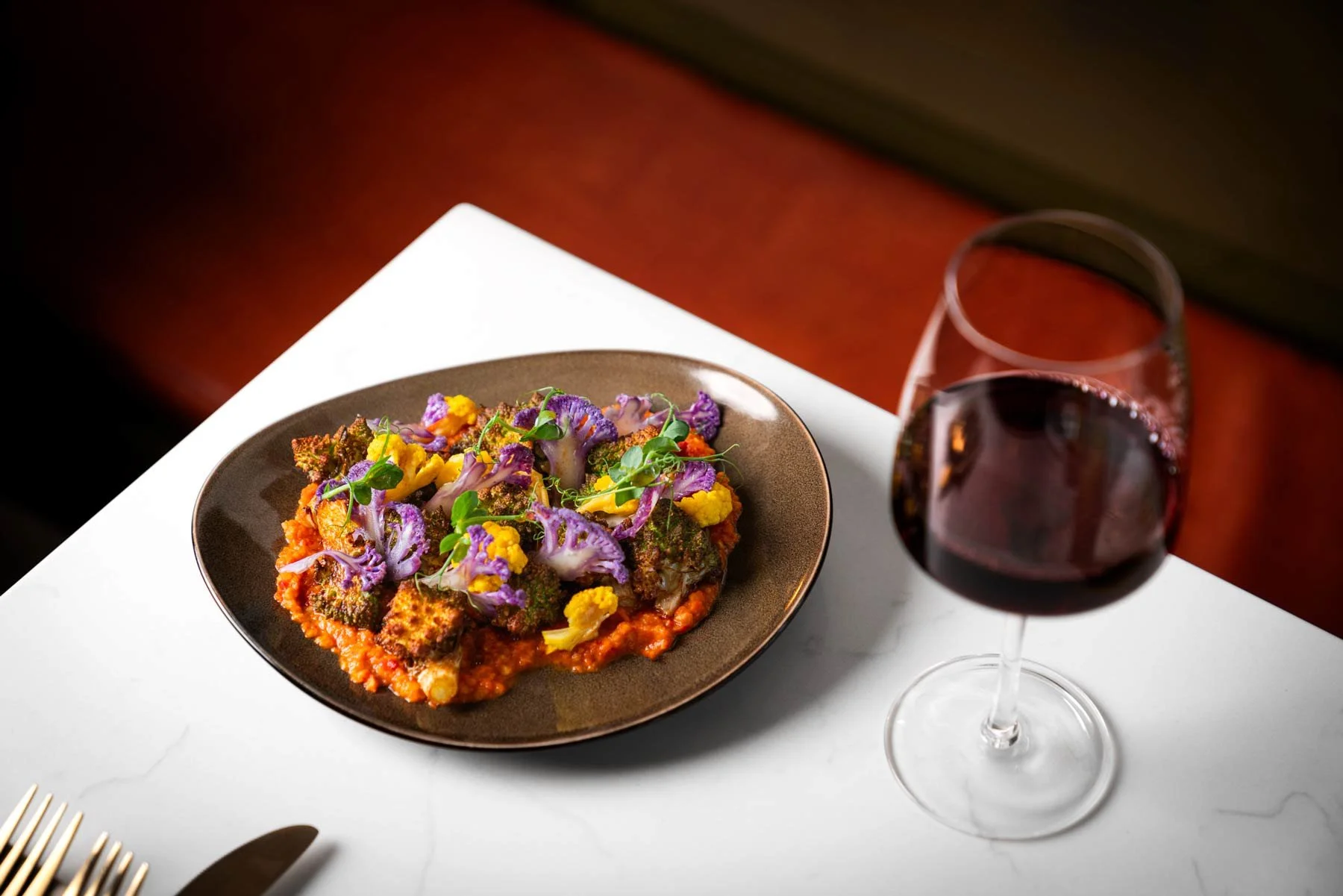 A plate of colorful roasted vegetables on a ceramic dish next to a glass of red wine on a white table.