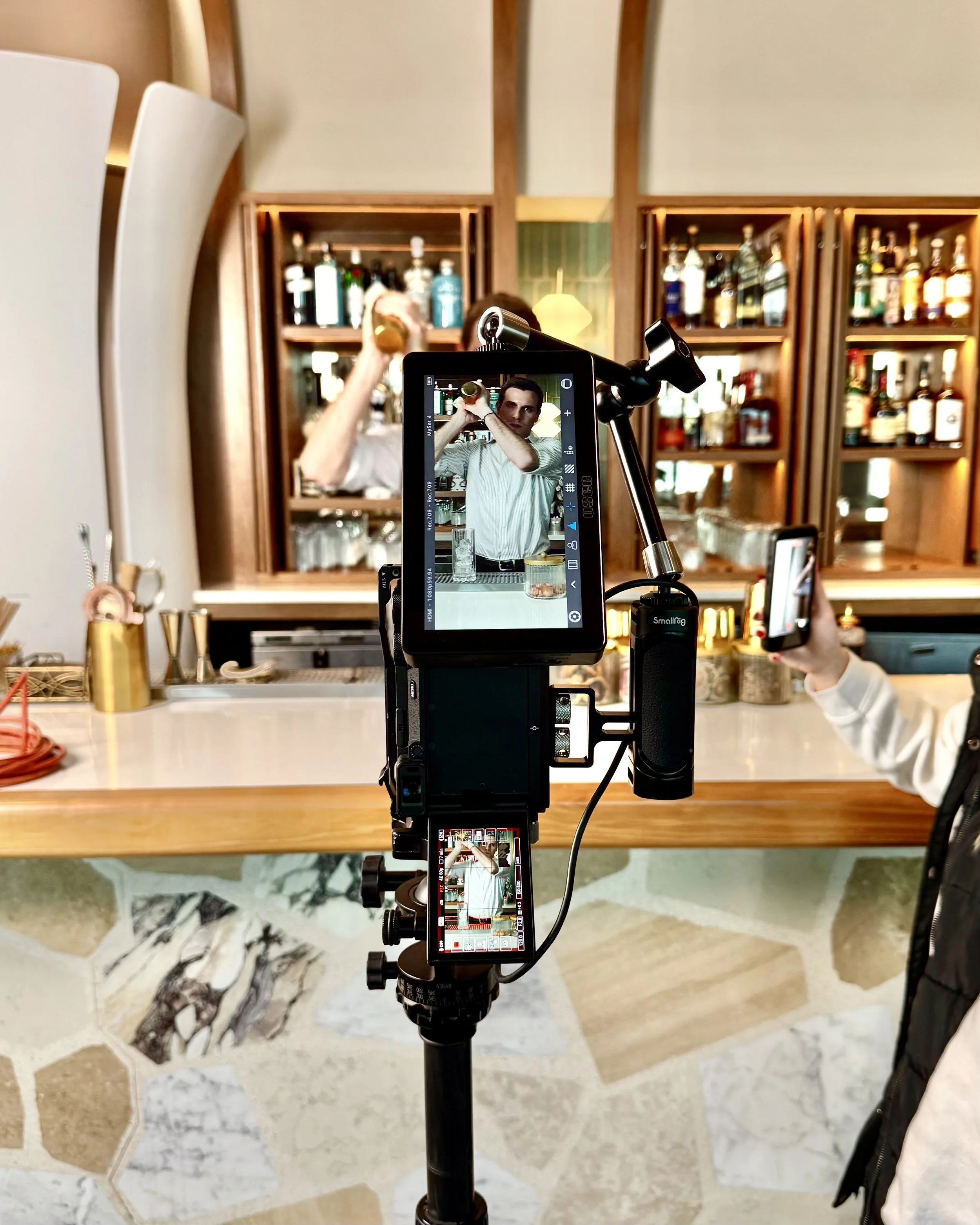A person filming a bartender pouring a drink behind a bar, with various bottles of alcohol displayed on shelves in the background.