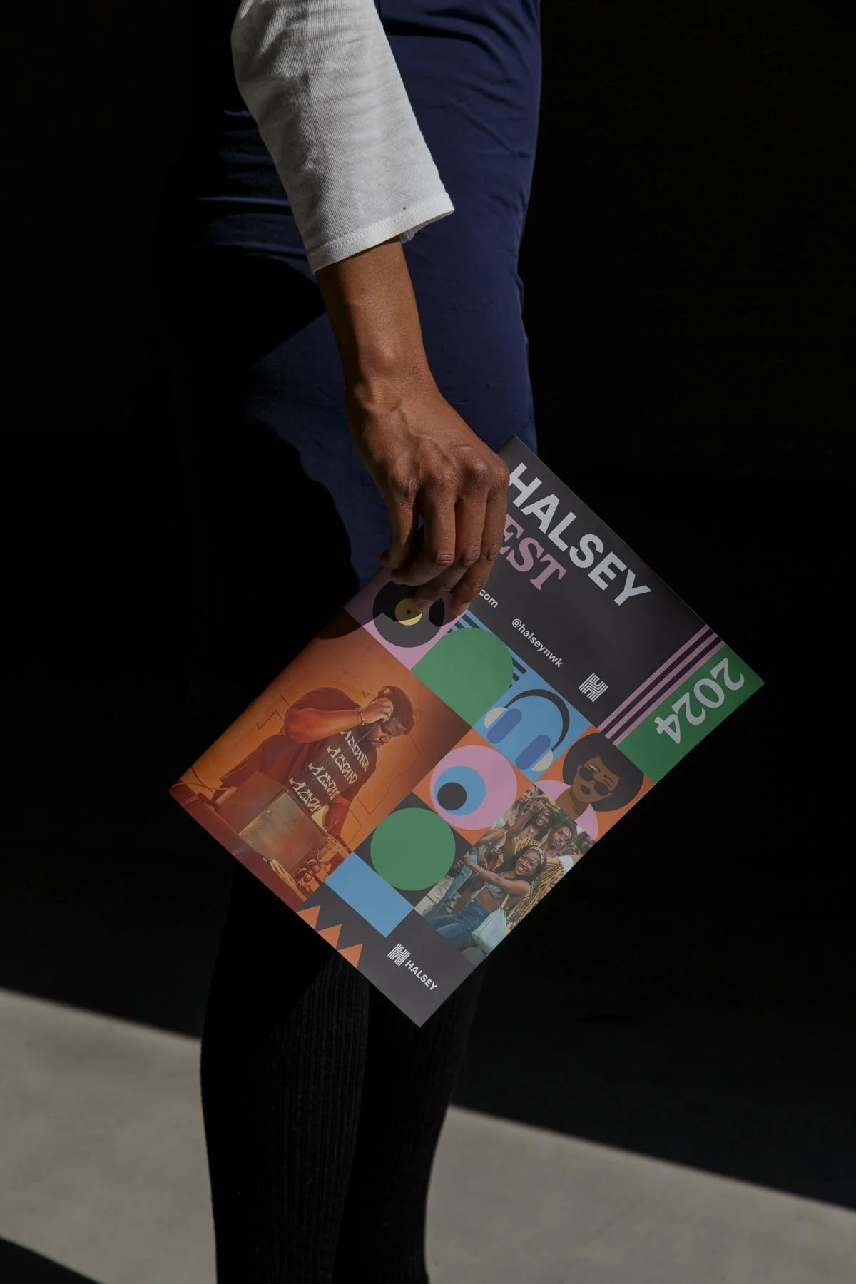 A person dressed in a white shirt and dark blue pants holding a colorful event flyer in their right hand, with a black background.
