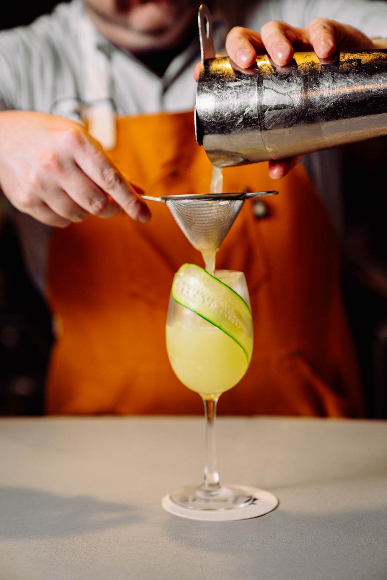 Bartender pouring a cocktail through a strainer into a glass garnished with a cucumber ribbon.