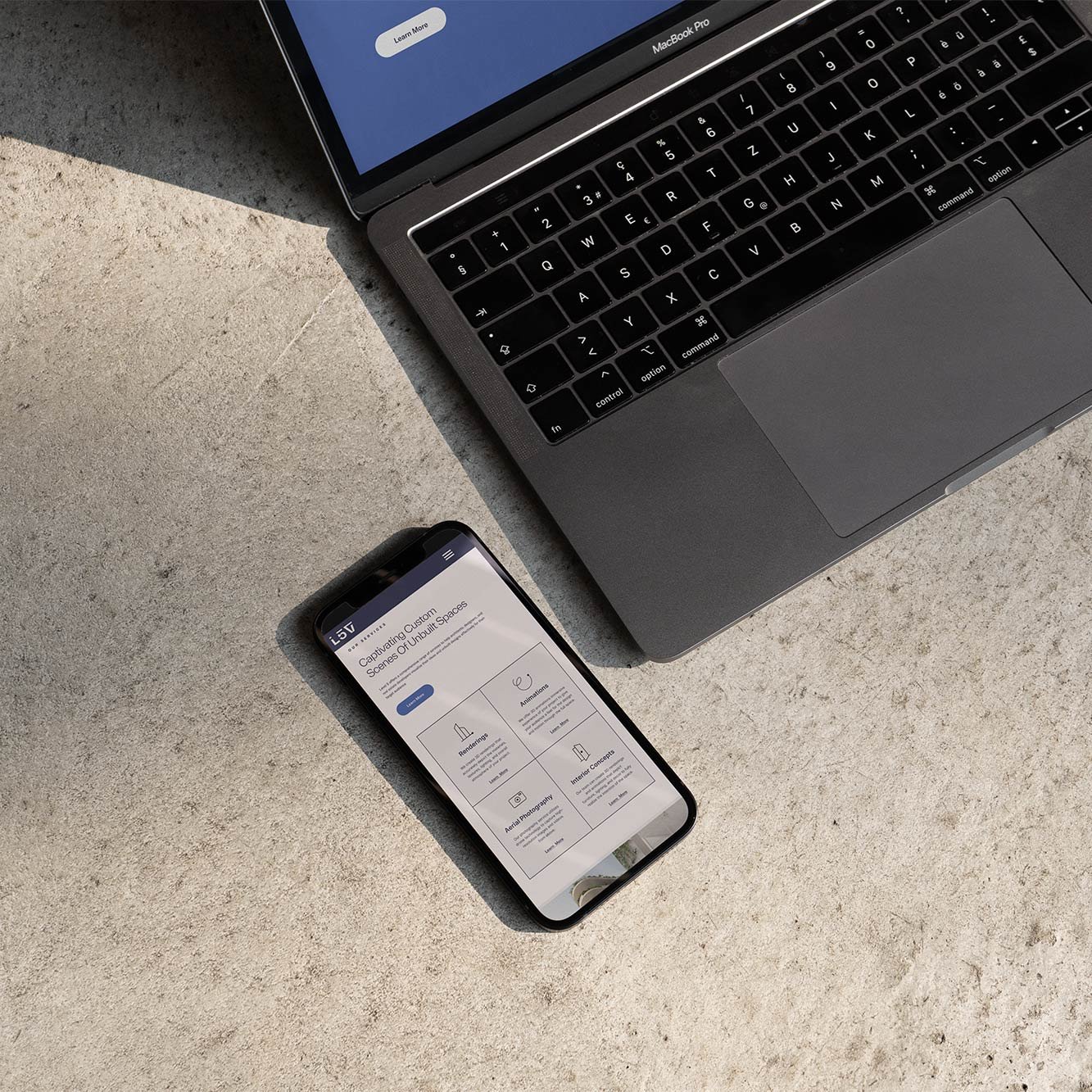 A laptop and a smartphone placed on a concrete surface, with the laptop showing a blue screen and the smartphone displaying a webpage about capturing custom scenes in a creative app.