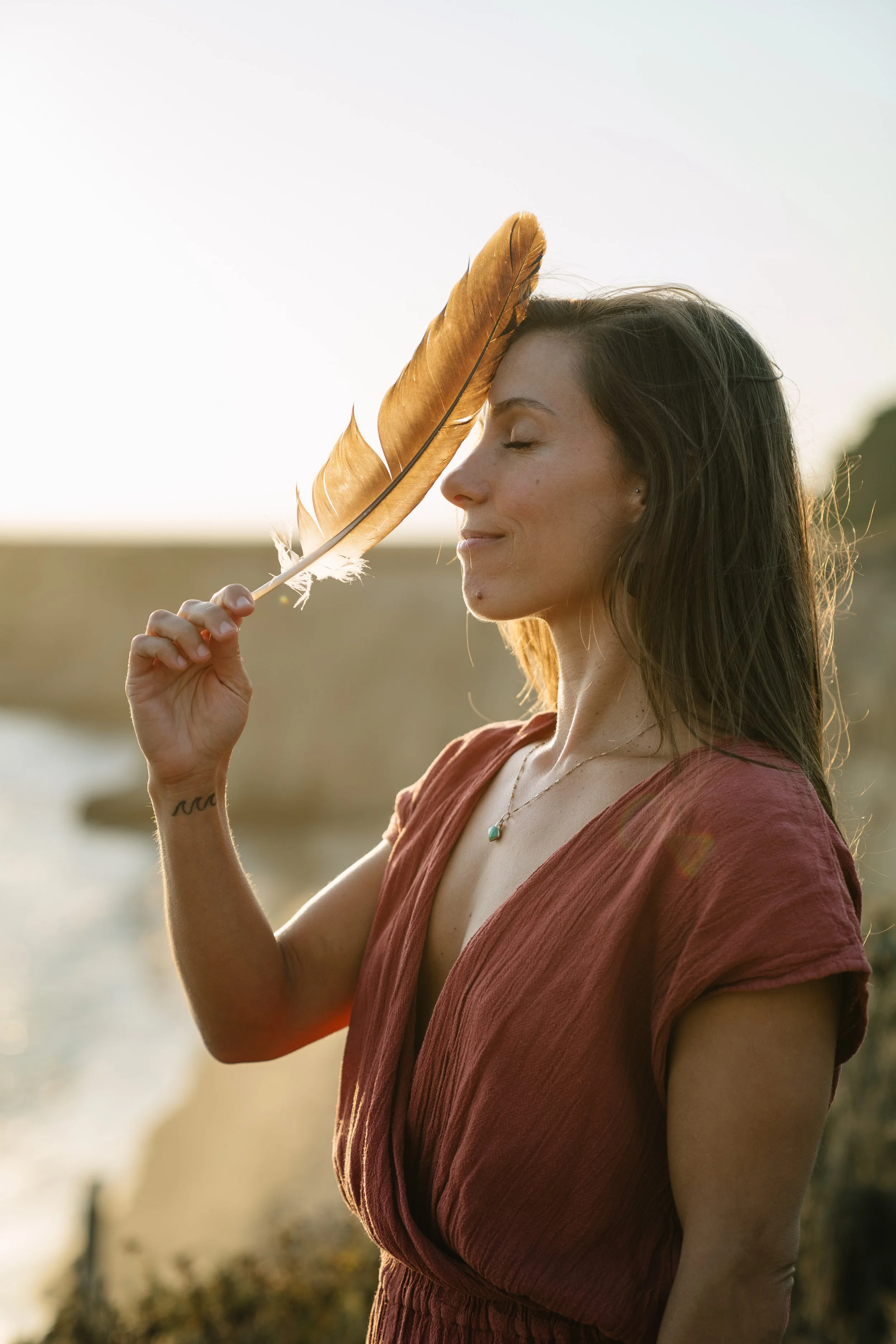 Abby on a coastal bluff at sunset, eyes closed, holding a large feather to her forehead