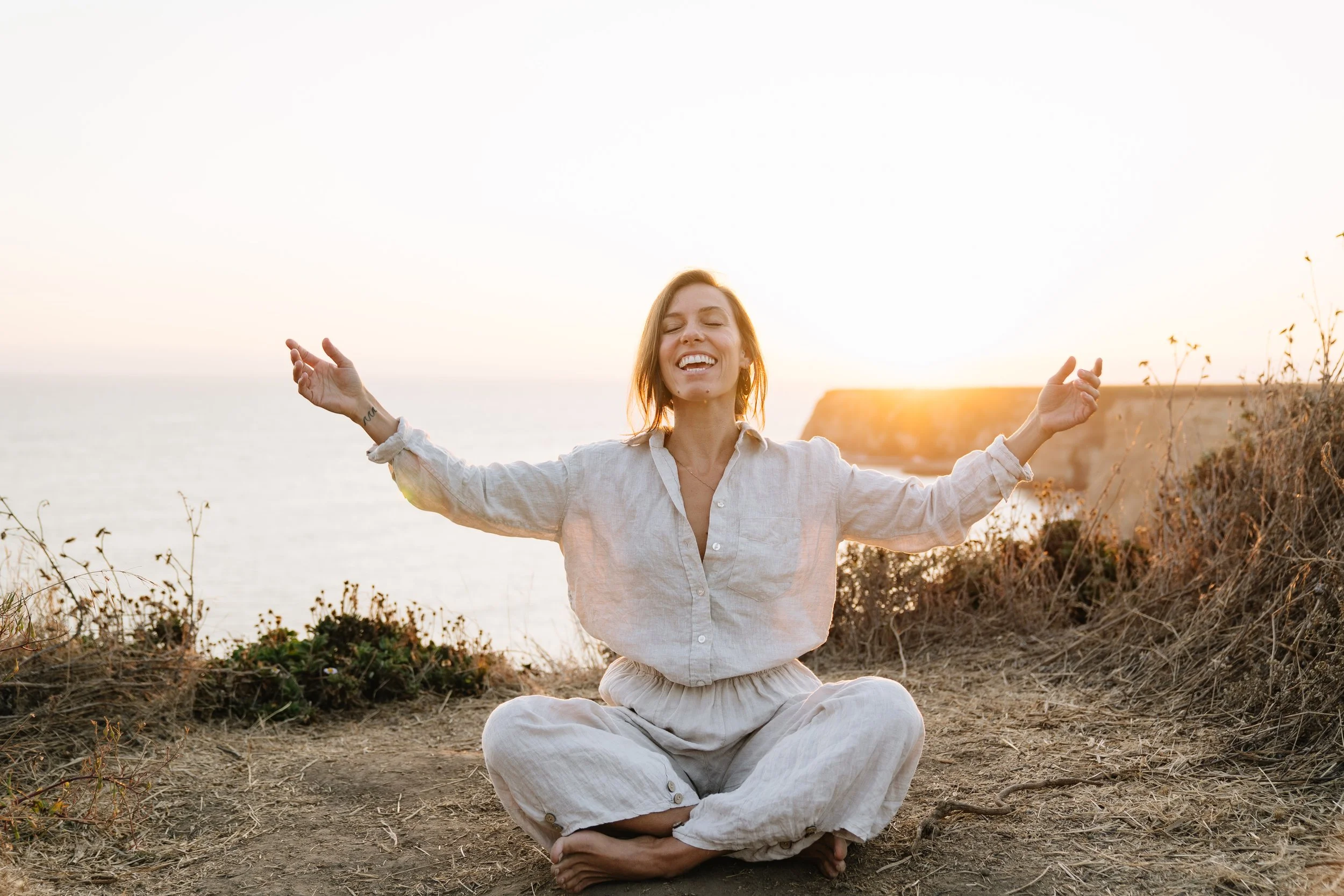 Abby sitting cross-legged on a bluff above the ocean at sunset, arms open, laughing