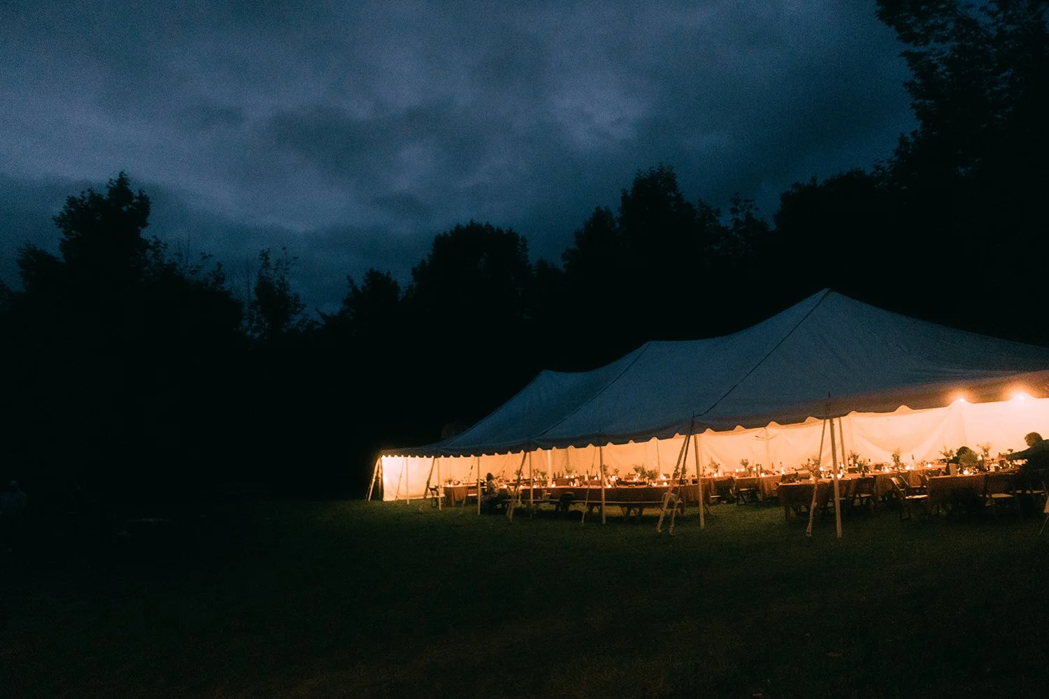 A large outdoor event tent illuminated from inside at dusk, set up on a grassy area with tables and chairs underneath, surrounded by trees and darkening sky.
