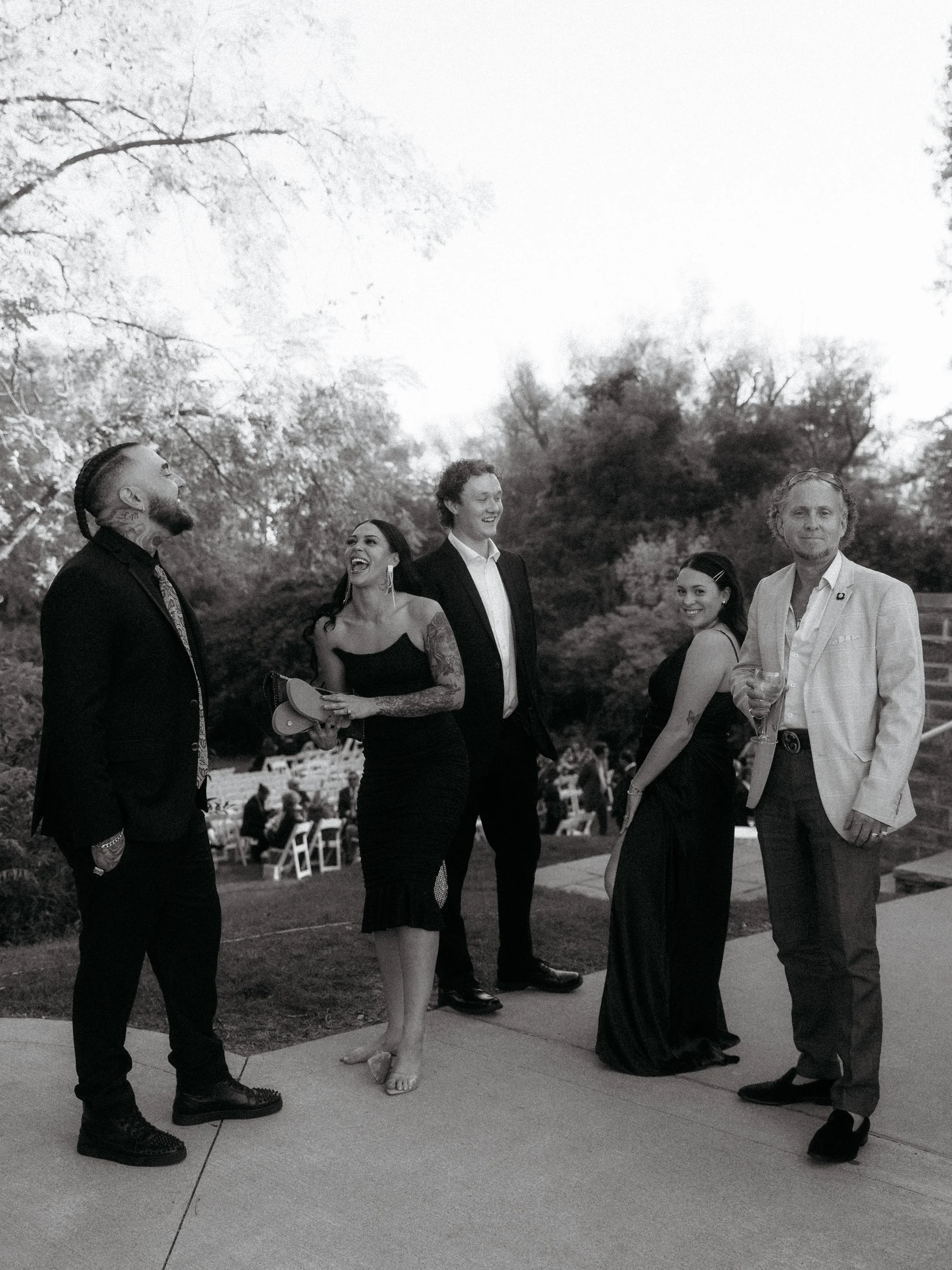 Group of five people dressed in formal attire standing outdoors, enjoying a social event with trees and chairs in the background.