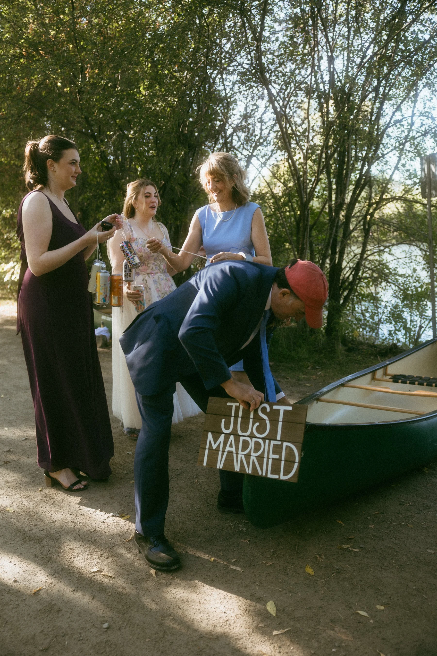 Group of people celebrating a wedding outdoors, with one person holding a 'Just Married' sign, near a kayak by a river, surrounded by trees.