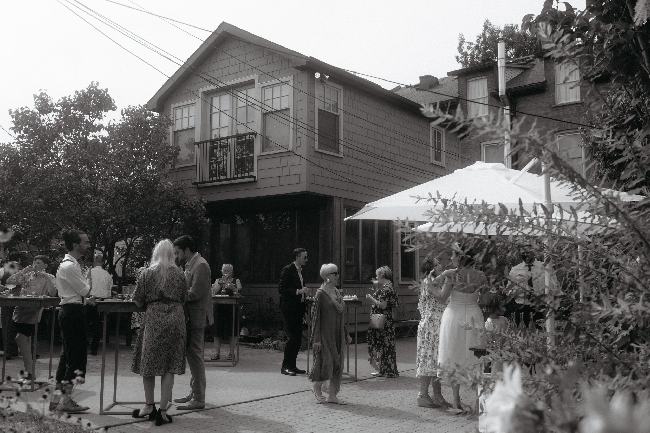 People socializing outdoors in a backyard, some holding drinks, with a house and trees in the background, and a large umbrella providing shade.