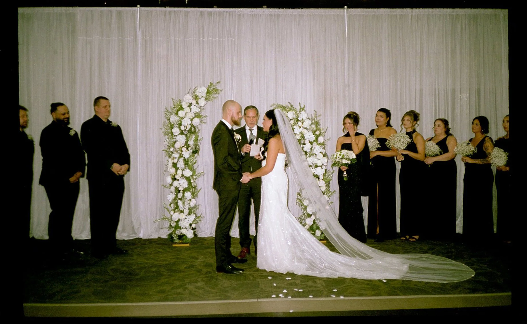 A wedding ceremony with a couple holding hands in front of an officiant, surrounded by bridesmaids and groomsmen, with floral arrangements as backdrop.