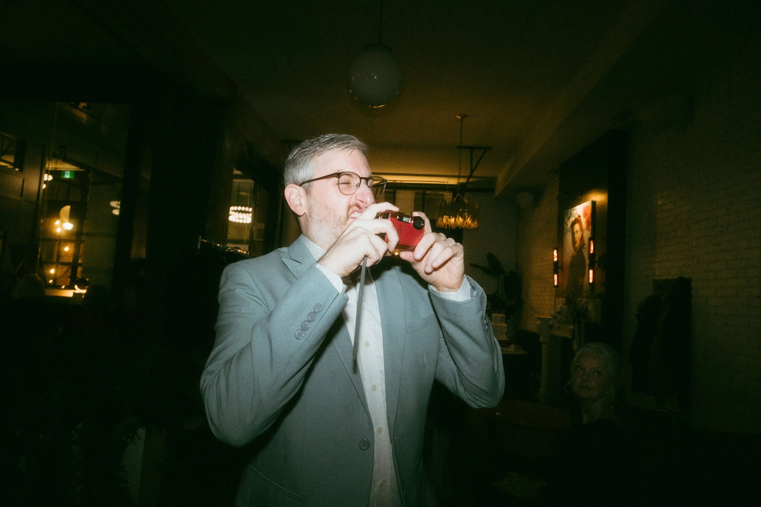 A man in a gray suit and glasses taking a photo with a red camera in a dimly lit indoor setting, with a statue of an elderly woman in the background.