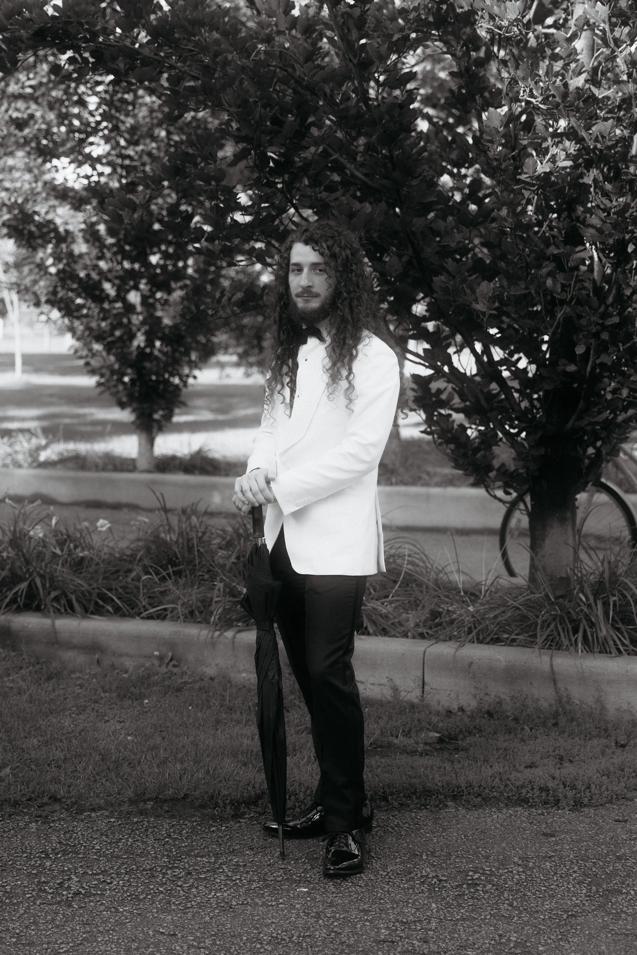 A man with long curly hair and a beard is standing outdoors in front of a tree, wearing a white blazer, black pants, and shiny shoes, holding a black umbrella.