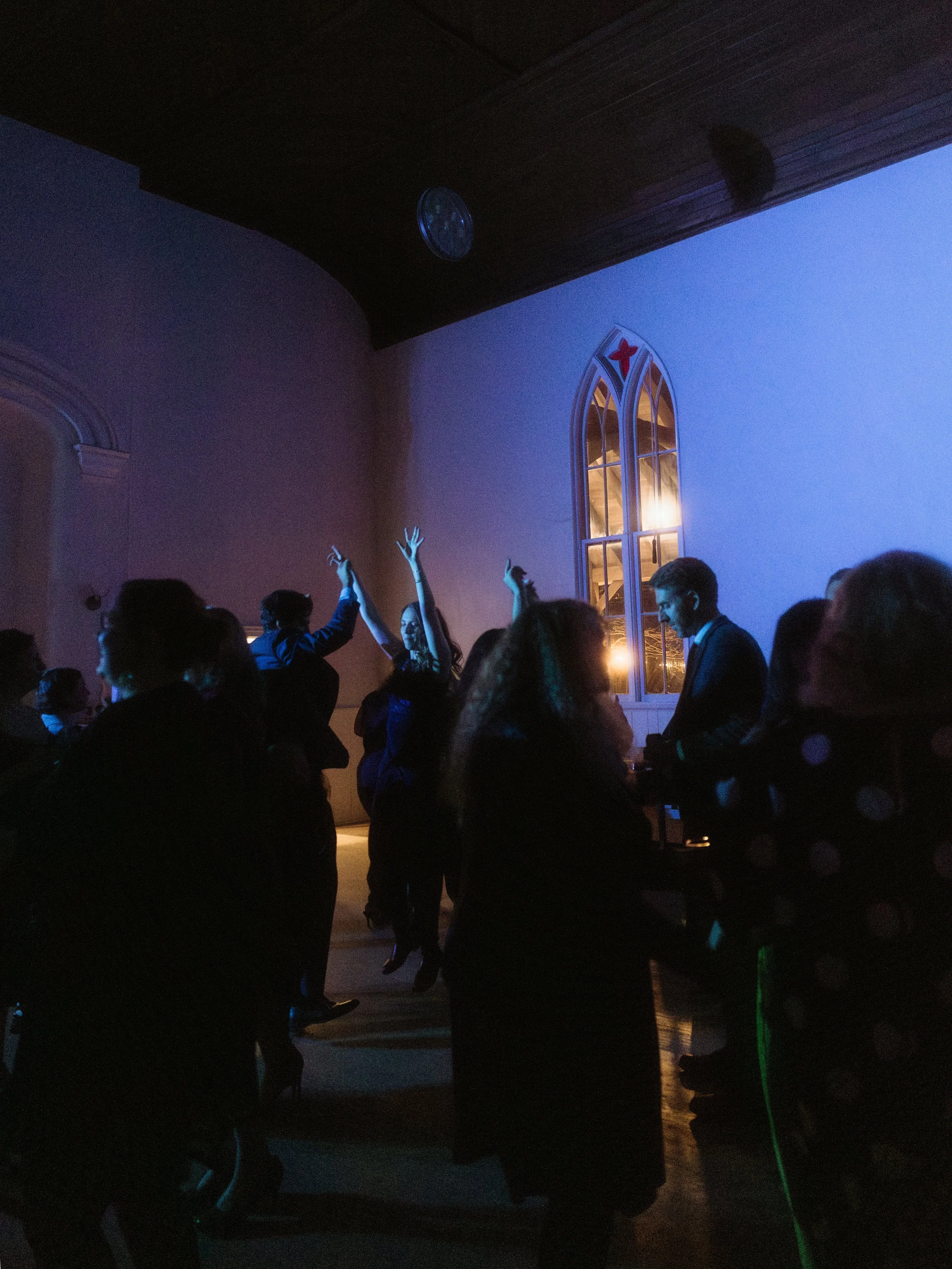 People dancing and socializing inside a dimly lit church with stained glass windows and a red cross symbol.