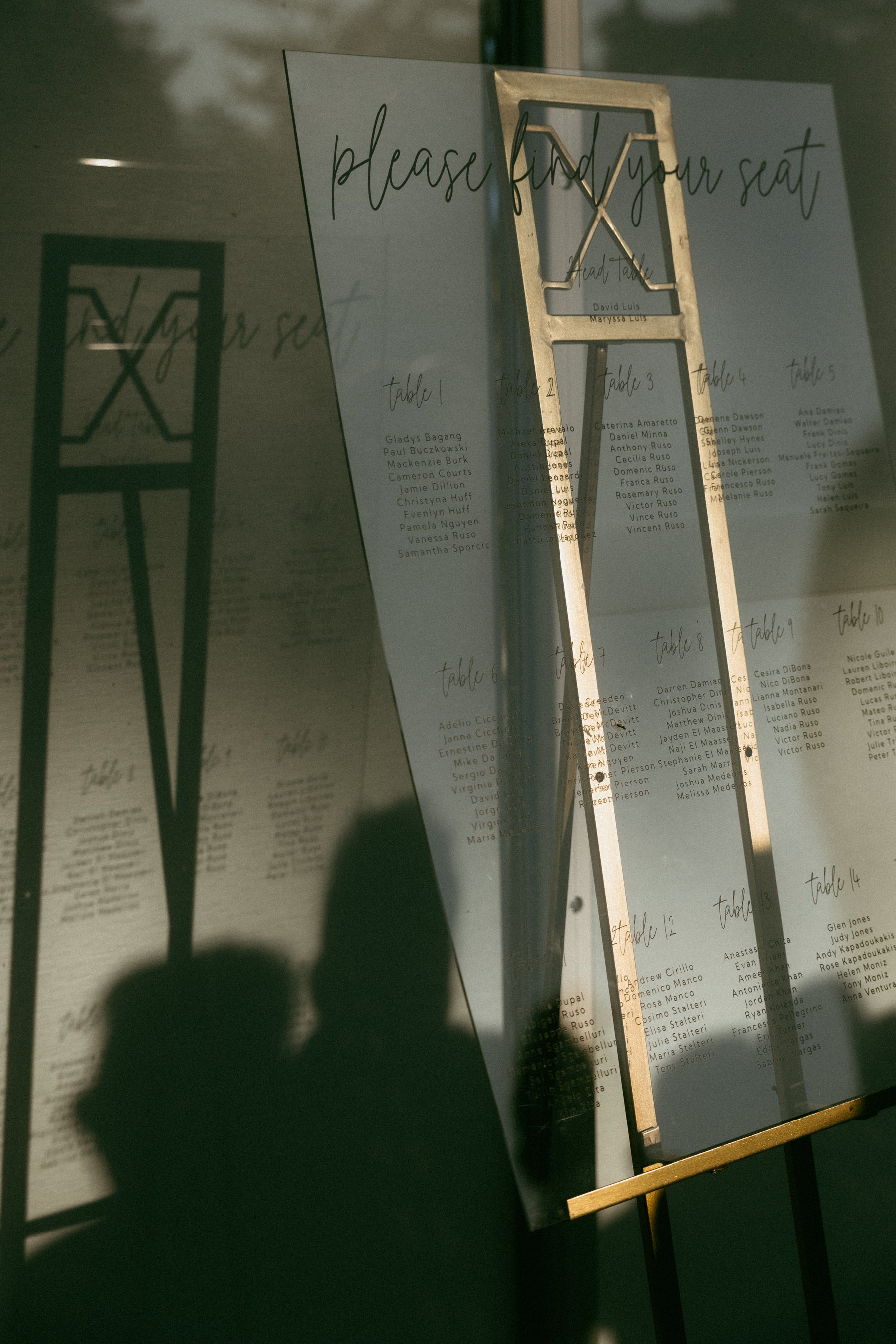 A sign asking guests to find their seats at an event, with a large X-shaped stand holding a transparent sheet with handwritten-style text listing table assignments. The sign reads 'please find your seat' and includes names of guests.