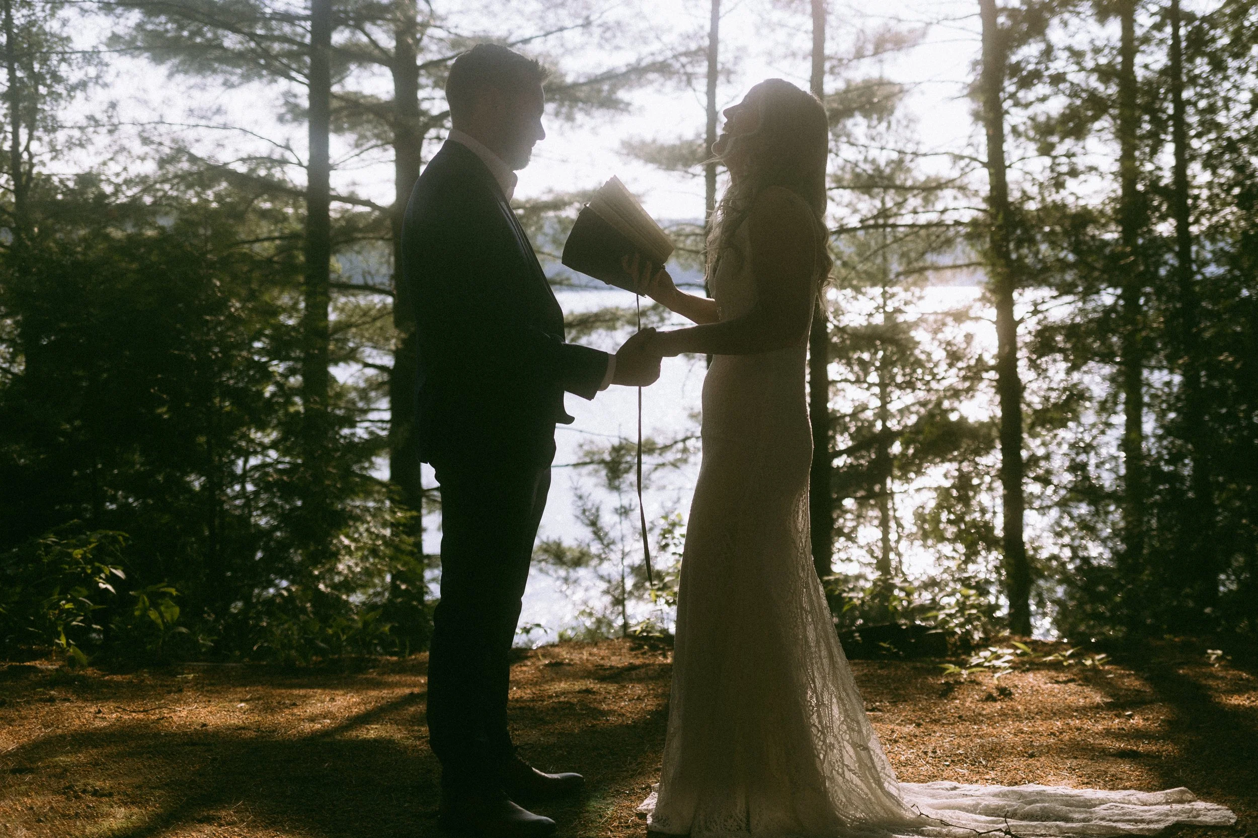 A couple getting married outdoors near a lake, holding hands, with the officiant reading from a book, surrounded by trees.