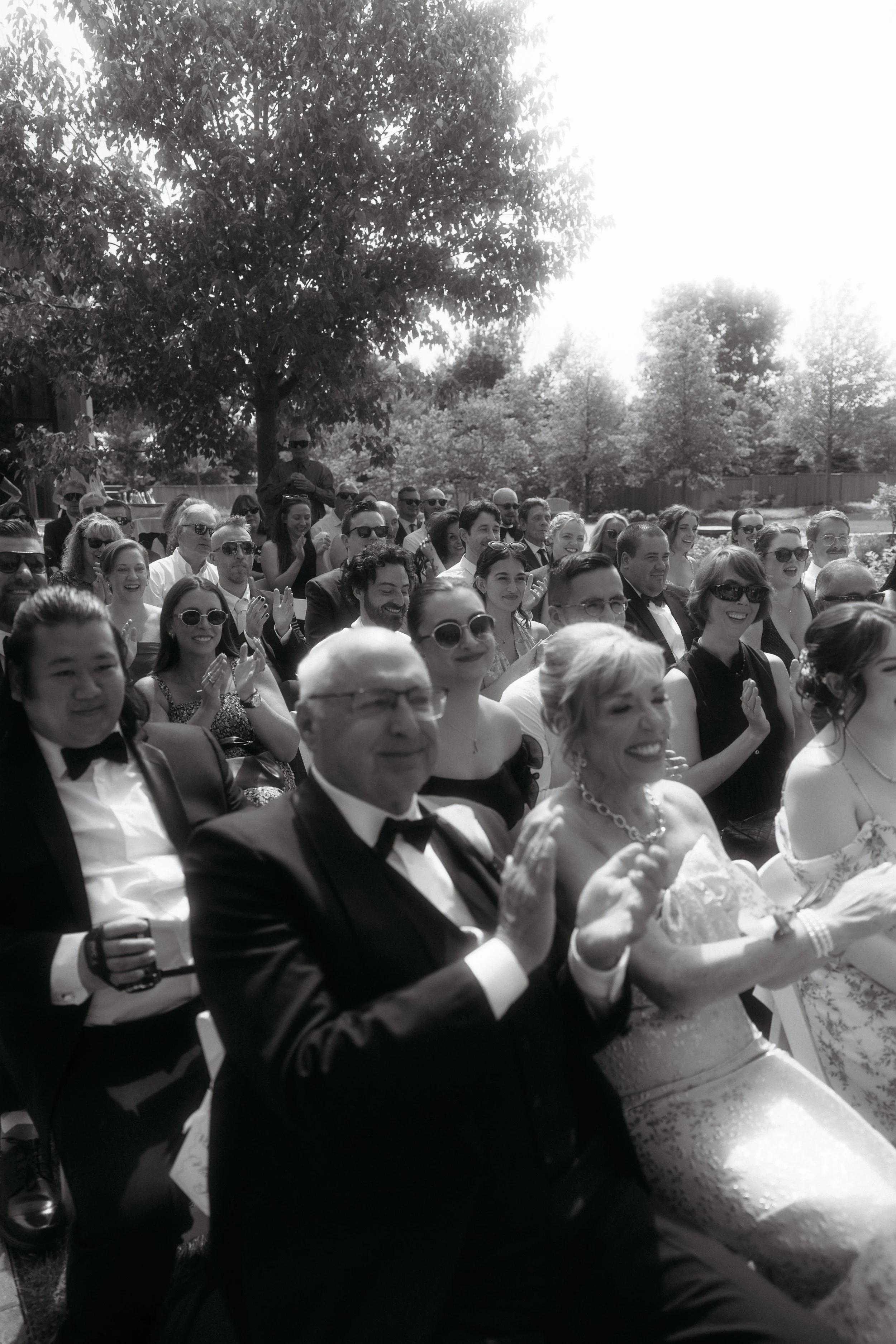 Black and white photo of a wedding crowd outdoors, seated on chairs, clapping, smiling, and enjoying the event around a large tree with sunlight filtering through the leaves.