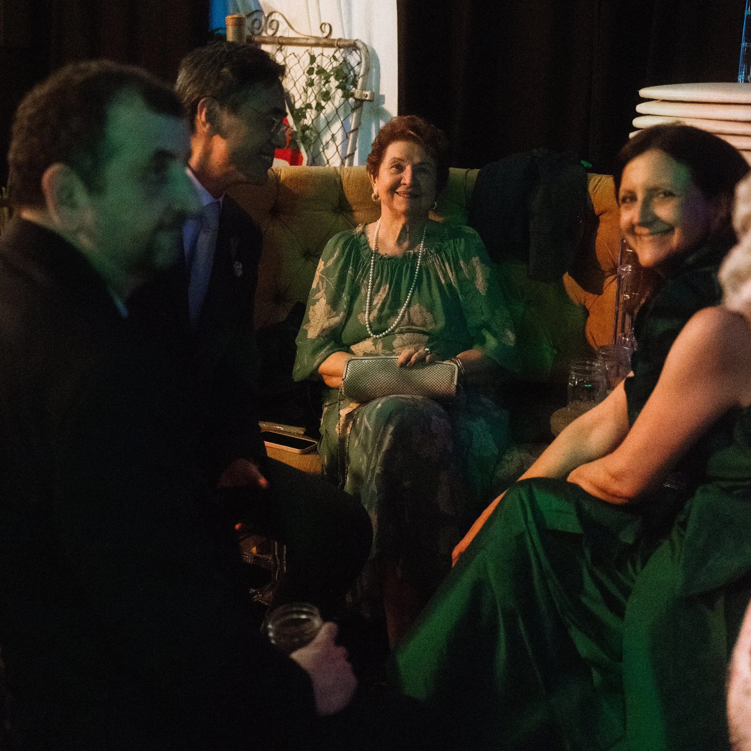 Group of five people sitting and talking in a dimly lit setting, with a woman in a green dress and pearl necklace at the center at Objx Studio in Toronto
