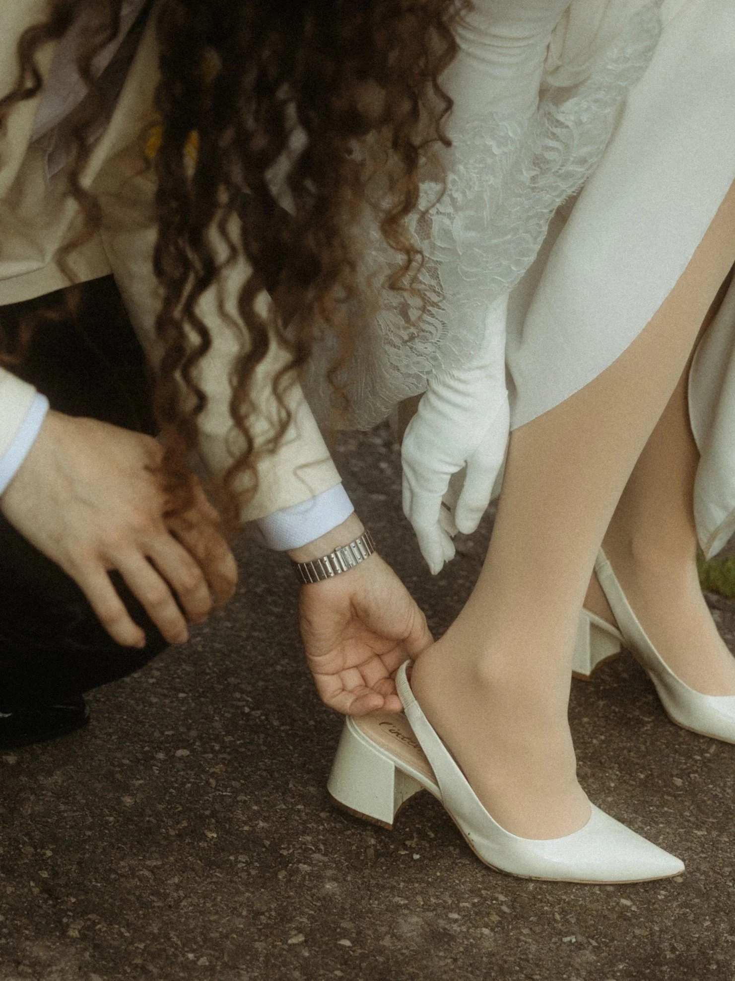 A person is helping a bride put on her cream-colored high heel shoe. The bride is wearing a white dress and cream-colored tights, while the person assisting is dressed in beige/cream attire with a watch.