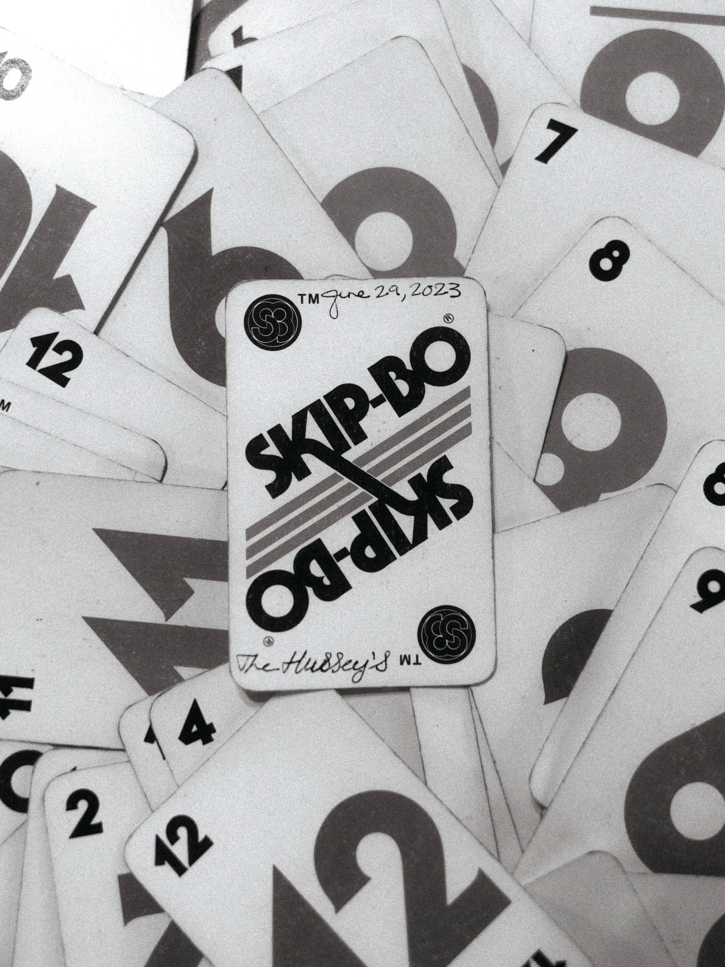 A black and white photograph of scattered Skip-Bo playing cards, with one card prominently displayed in the center showing the date 'June 29, 2023' and the name 'The Hudsey's' handwritten on it.