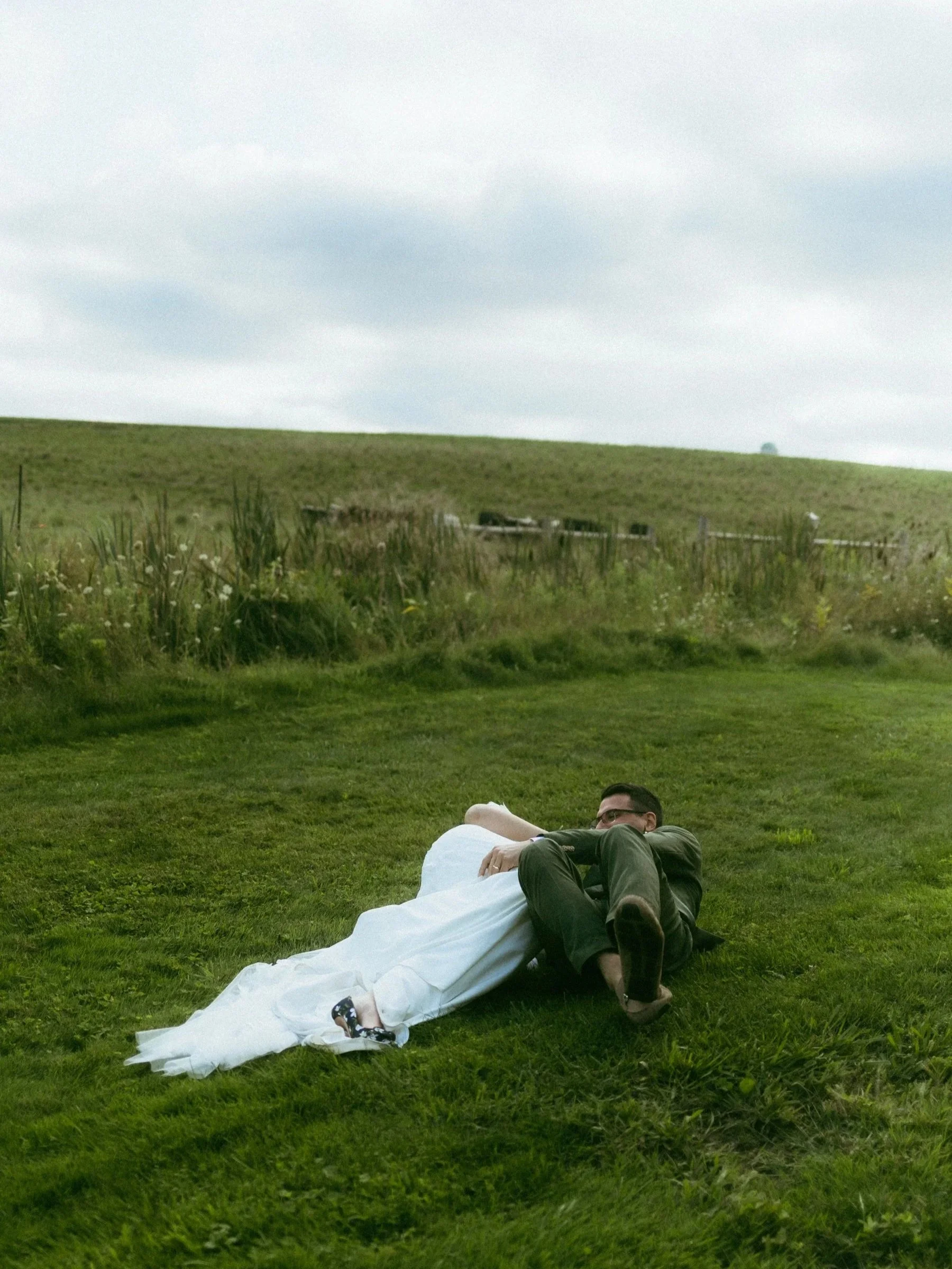 A man lying on the grass hugging a woman in a white wedding dress in an open field under a cloudy sky.