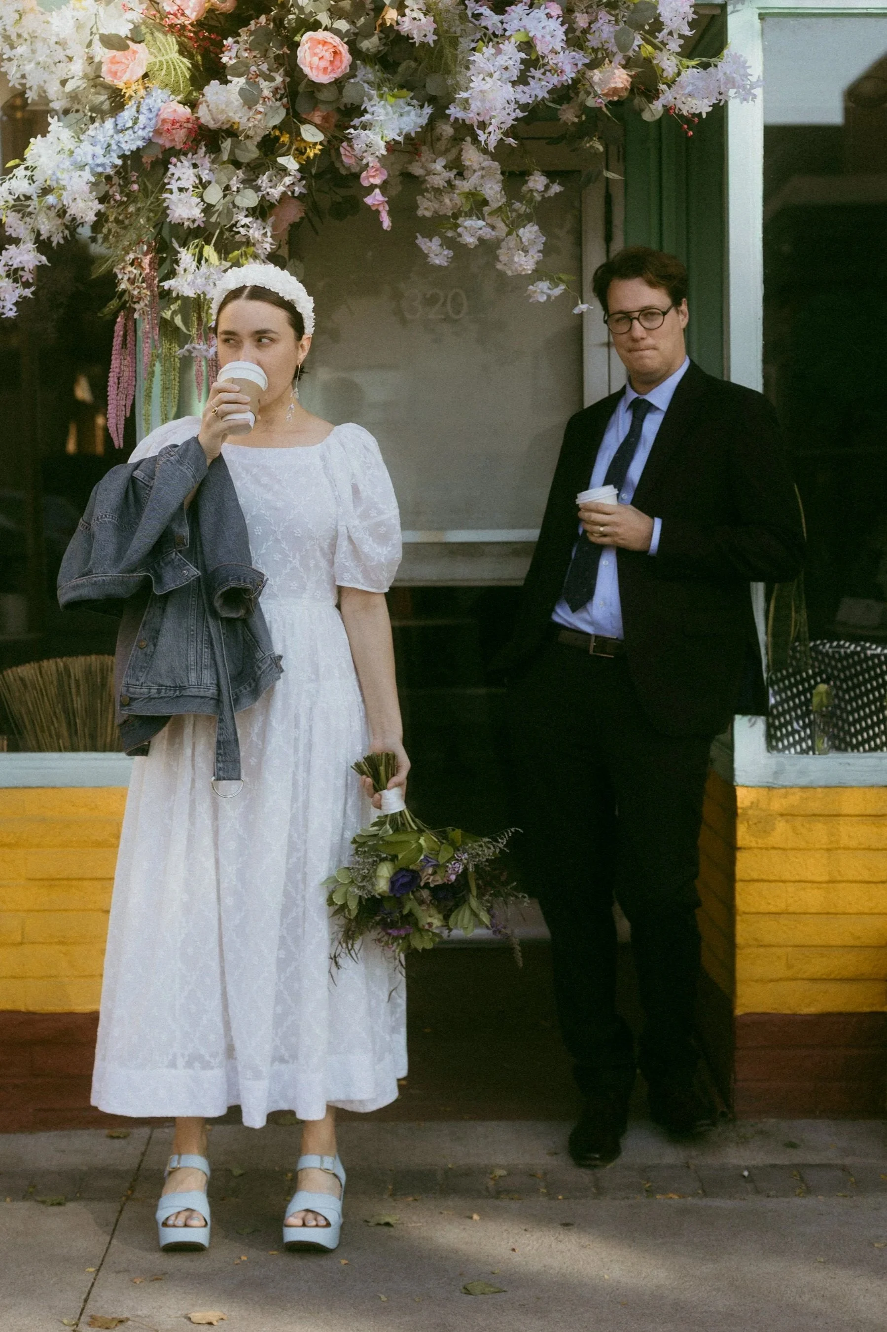 A woman in a white dress and platforms holding a bouquet of flowers, standing outside a floral decorated shop with a man in a dark suit and glasses near her, both holding coffee cups.