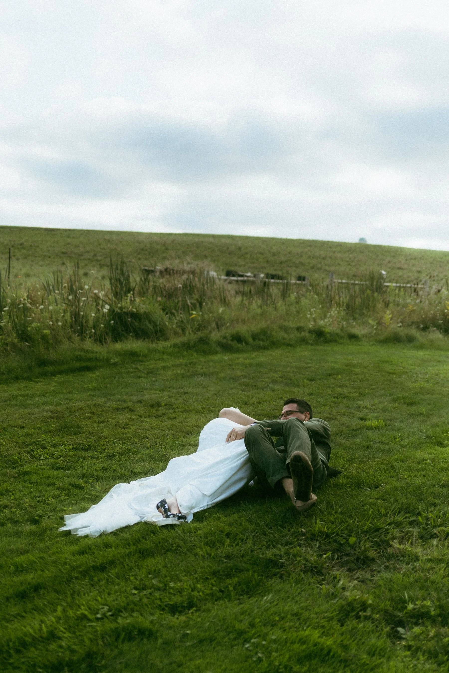 A bride and groom lying on grass, outdoors in a green field under a cloudy sky on their wedding day at Elle by Stella in St.Jacobs, Ontario